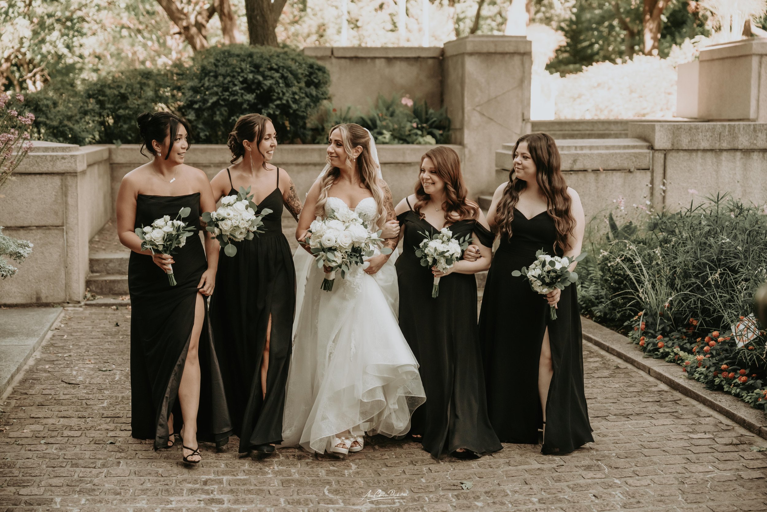A bride and five bridesmaids walking together outdoors, holding bouquets of white flowers, with greenery and stone steps in the background.