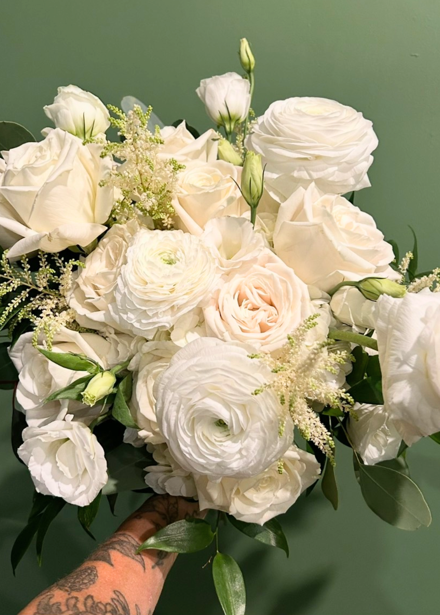 A bouquet of white and pale pink roses and ranunculus, with greenery and small white filler flowers, against a green background.