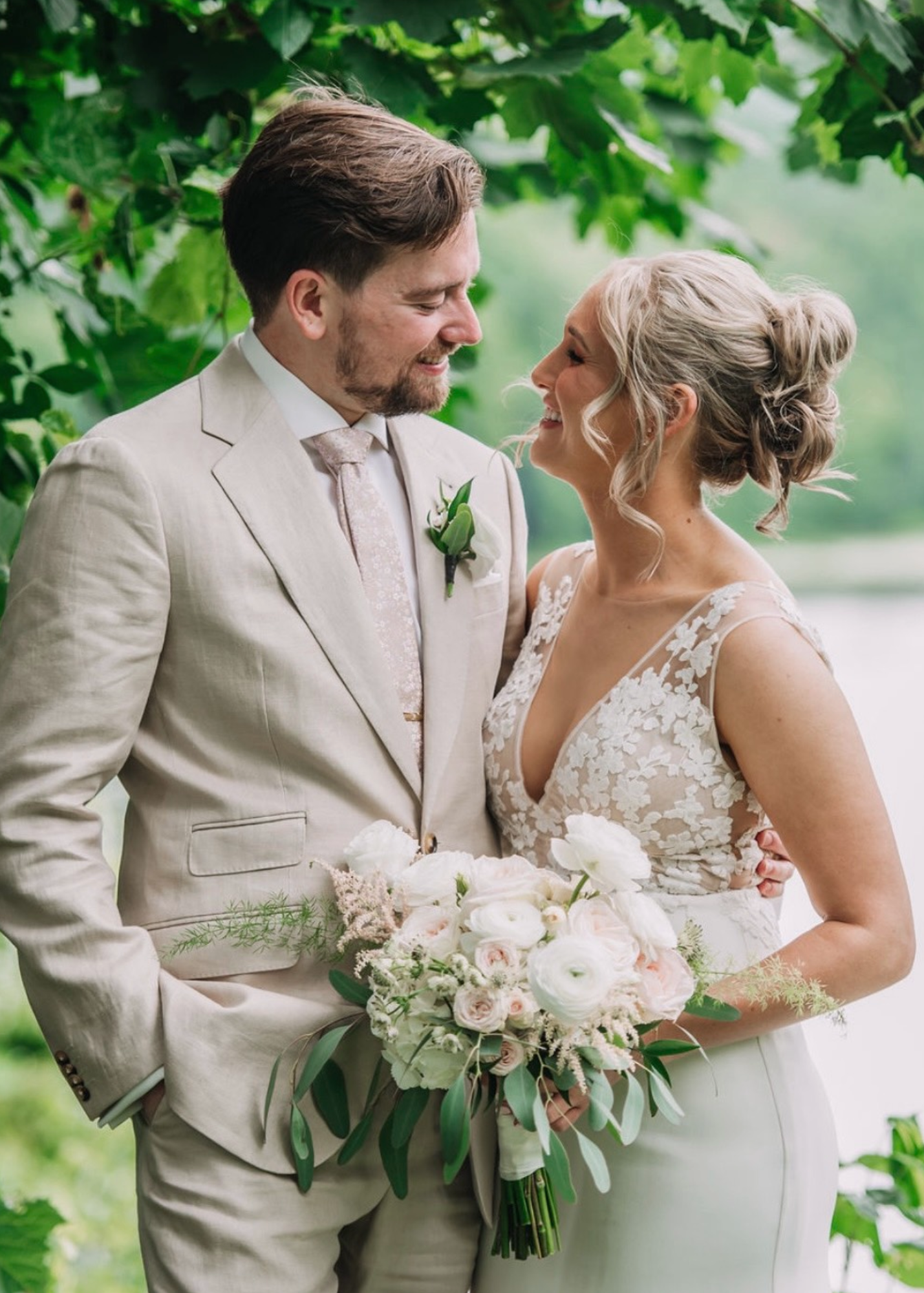 Bride and groom smiling and looking at each other outdoors, with greenery in the background. The bride holds a bouquet of white and pale pink flowers, wearing a lace dress, and the groom wears a light-colored suit with a boutonniere.