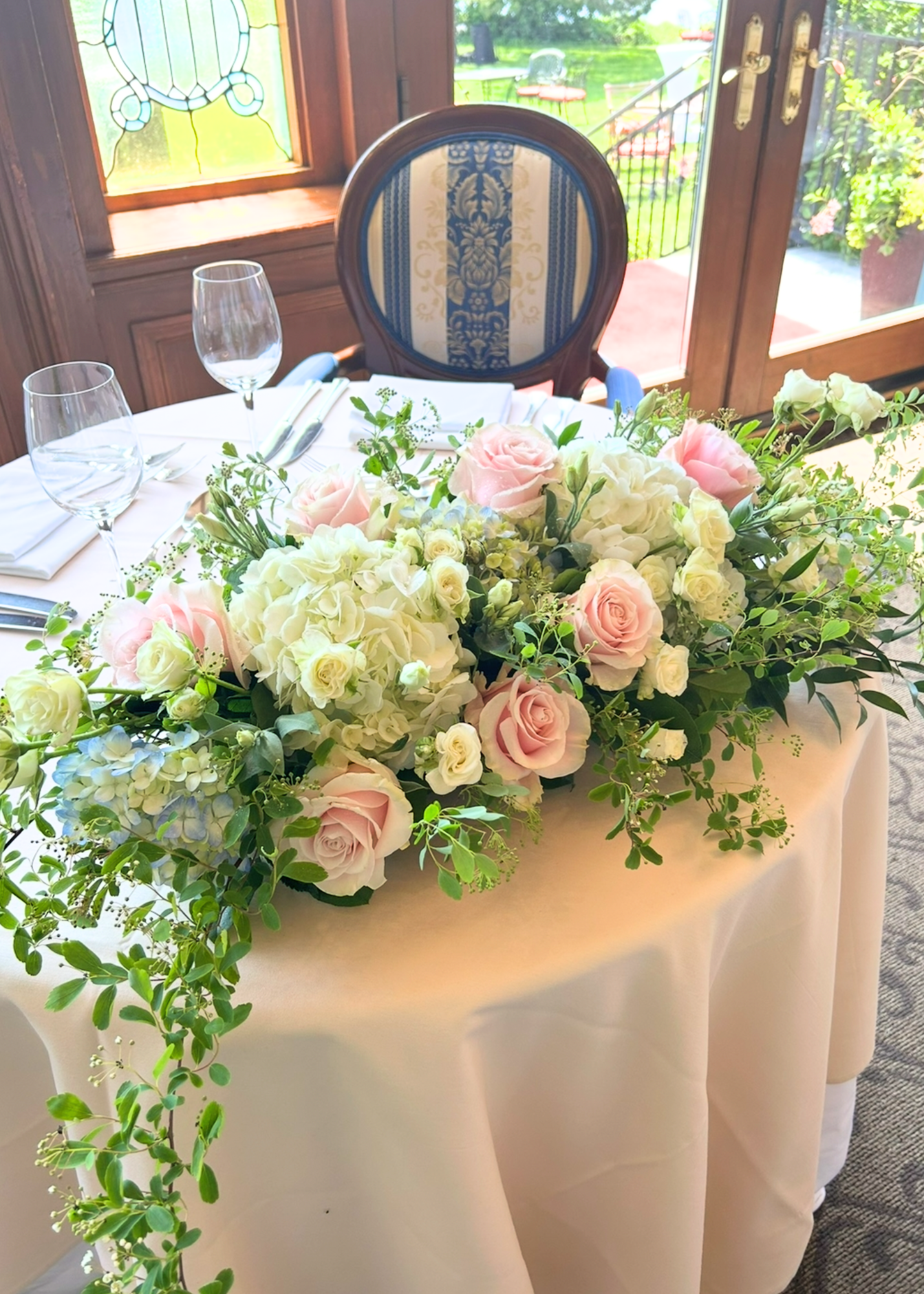 A round table with a black chair behind it, set for a formal meal with wine glasses, napkins, and silverware. The table features a large floral centerpiece with pink roses, white hydrangeas, and green foliage, in a bright room with wooden windows and a view of a patio and green outdoor space.