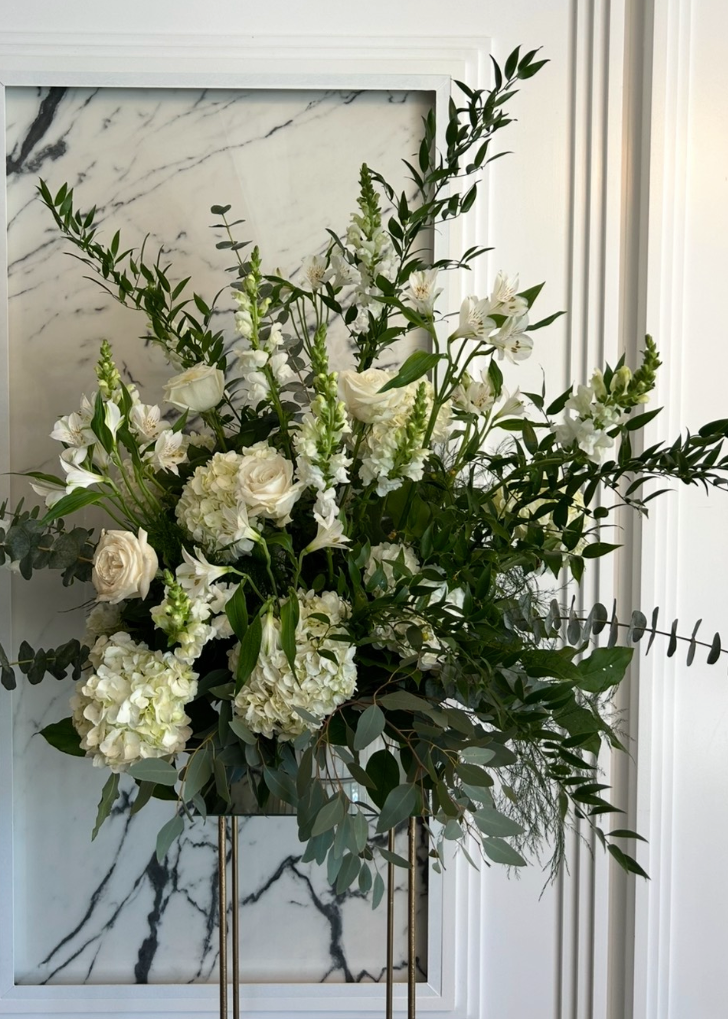 A large bouquet of white flowers and green foliage in a tall, metallic vase beside a marble wall.