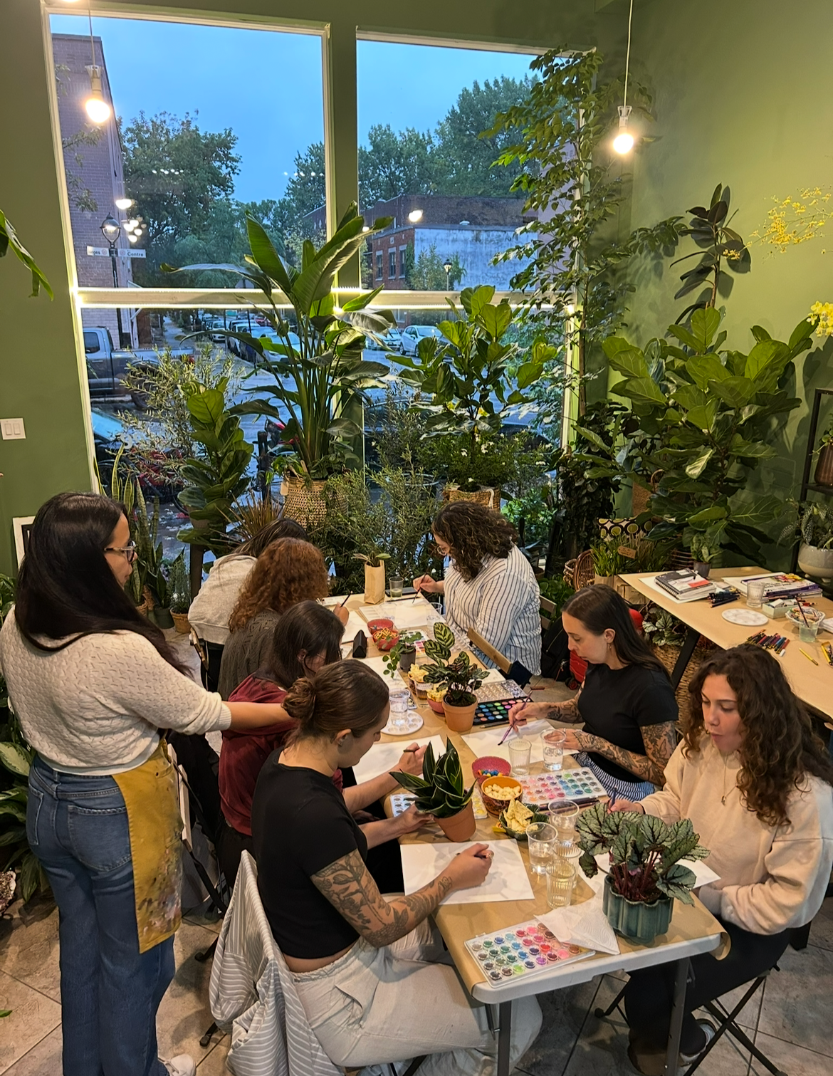People sitting around a table painting and drawing with various art supplies in a brightly lit room filled with large green houseplants, with a large window revealing an outdoor street scene.