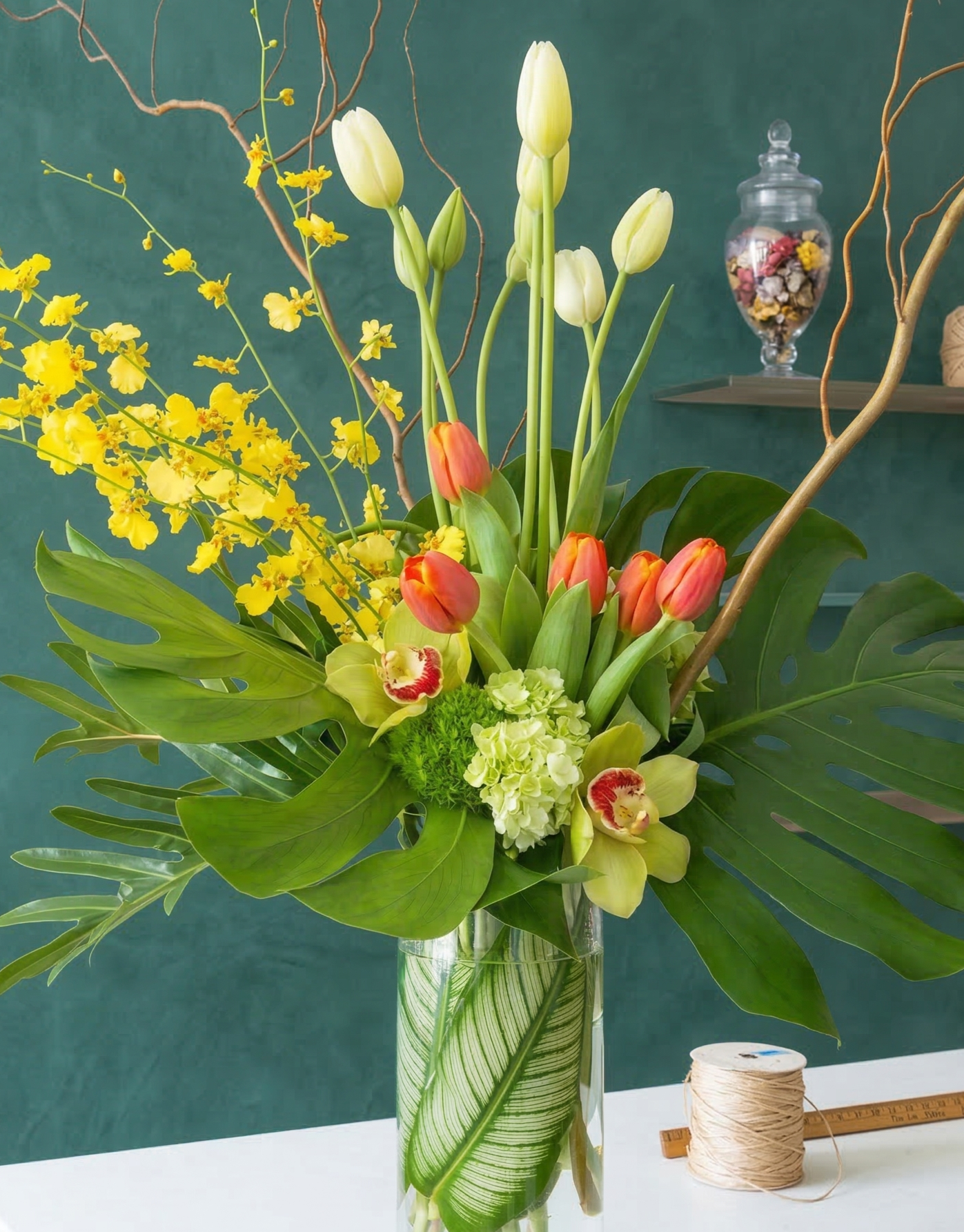 A vibrant floral arrangement in a glass vase featuring yellow, orange, and white flowers with green leaves, set against a dark green background.