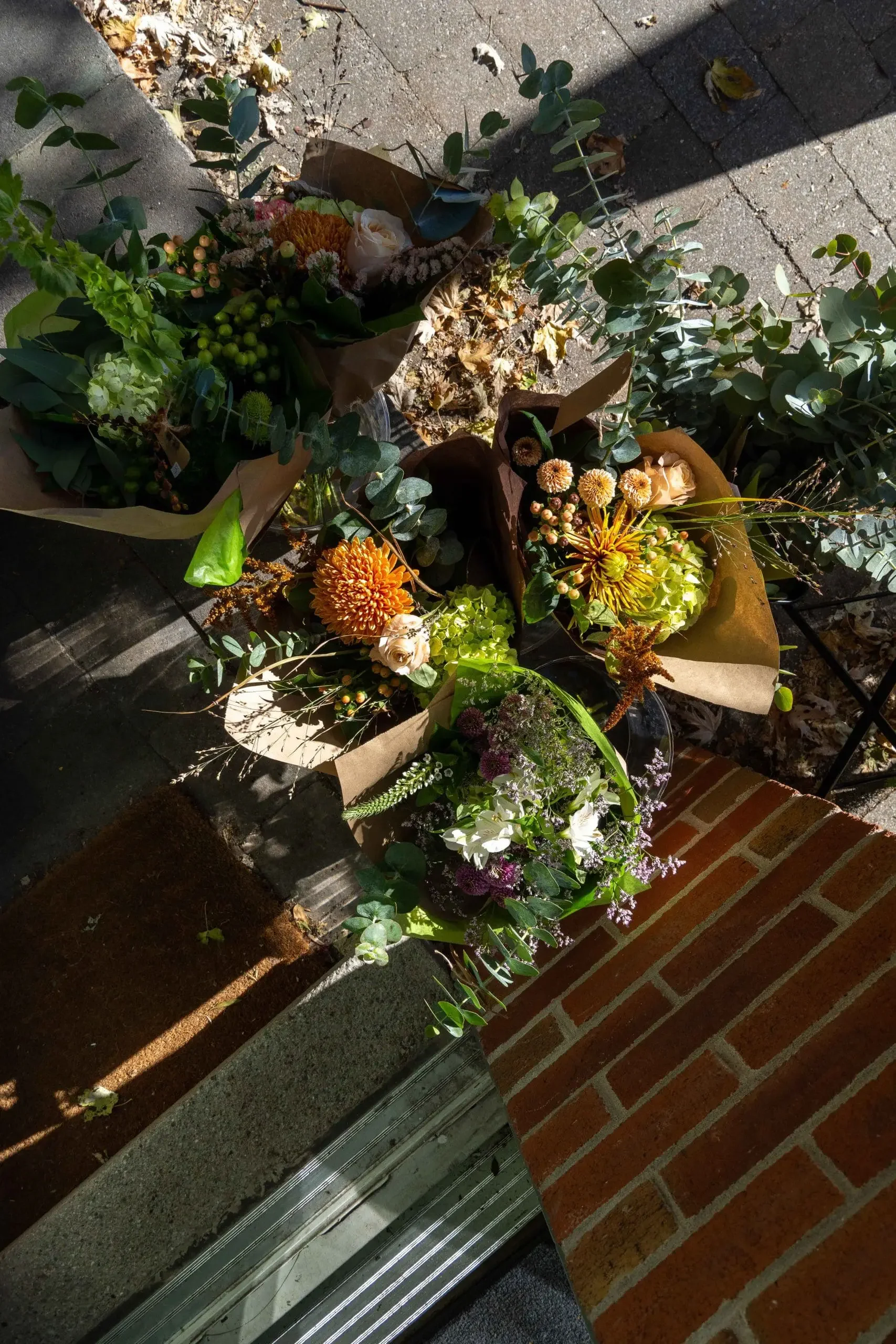 Several bouquets of flowers and greenery on a brick porch, illuminated by sunlight.