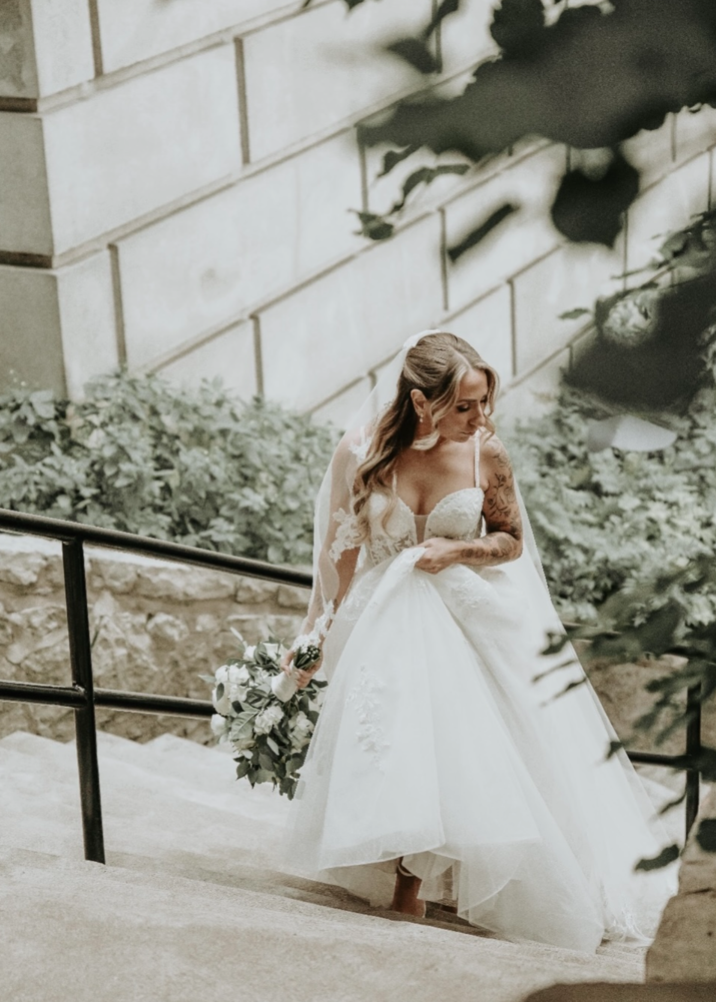 A bride in a white wedding dress holding a bouquet walks down a staircase outdoors, with stone walls and greenery around.