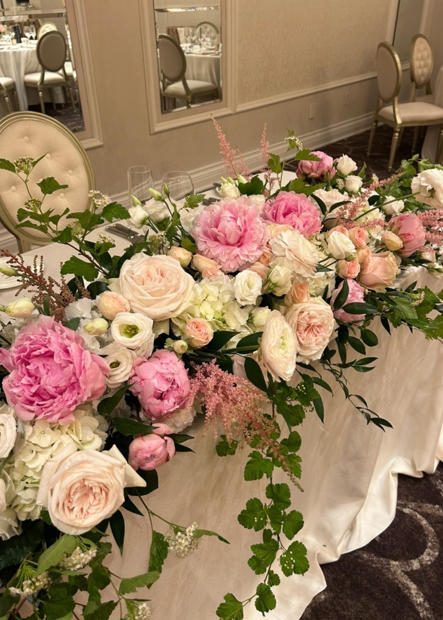 A floral centerpiece with pink, white, and blush roses, peonies, and greenery on a table for a special event or wedding reception.