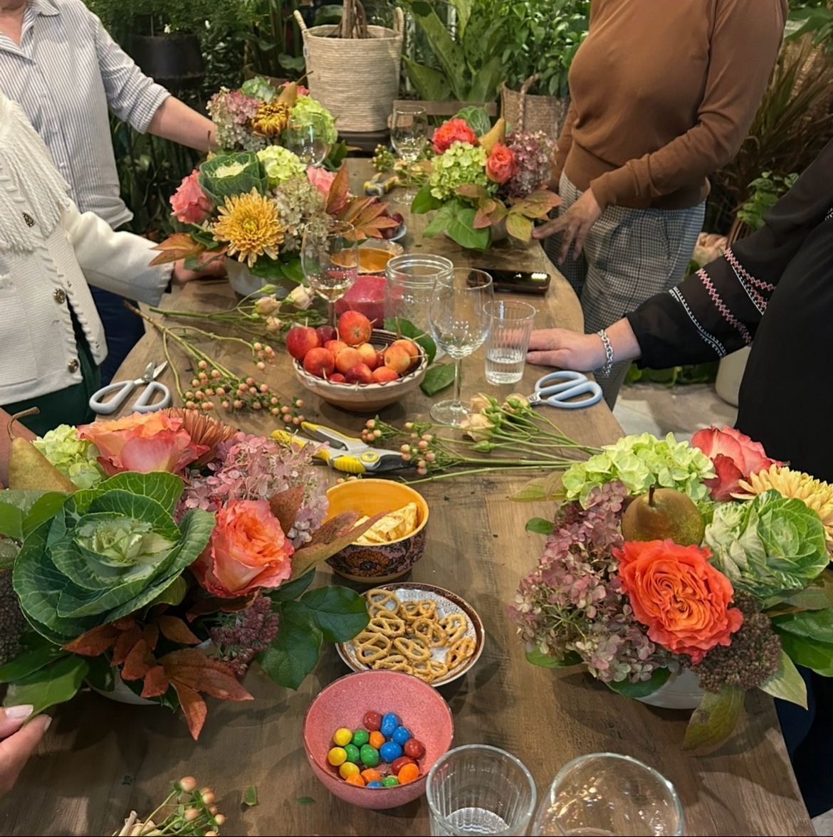 A gathering of people around a wooden table decorated with colorful flower arrangements, bowls of fruit, candy, pretzels, scissors, and cups of beverages in a lush greenery setting.