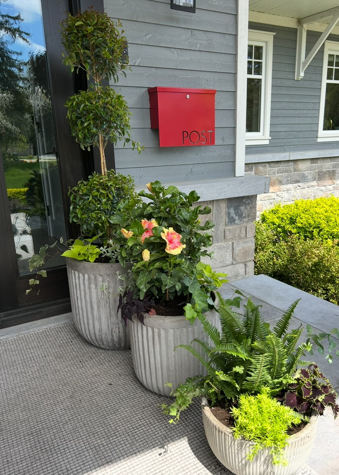 A red mailbox labeled 'POST' mounted on a blue wooden house exterior wall, with three large grey planter pots filled with various green plants and flowers in front.