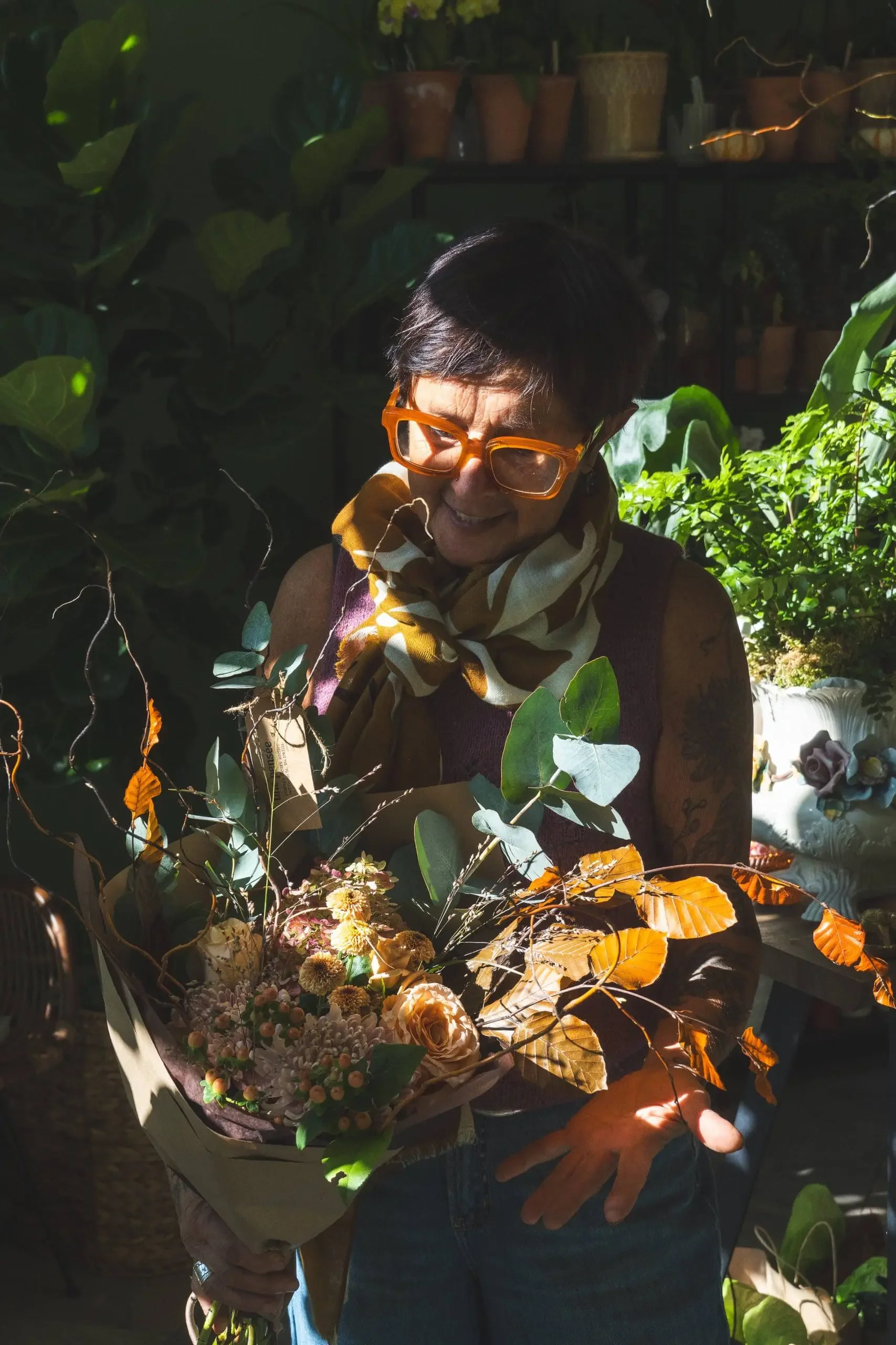 A woman wearing orange glasses and a patterned scarf is smiling while holding a bouquet of flowers and branches in a sunlit, plant-filled environment.