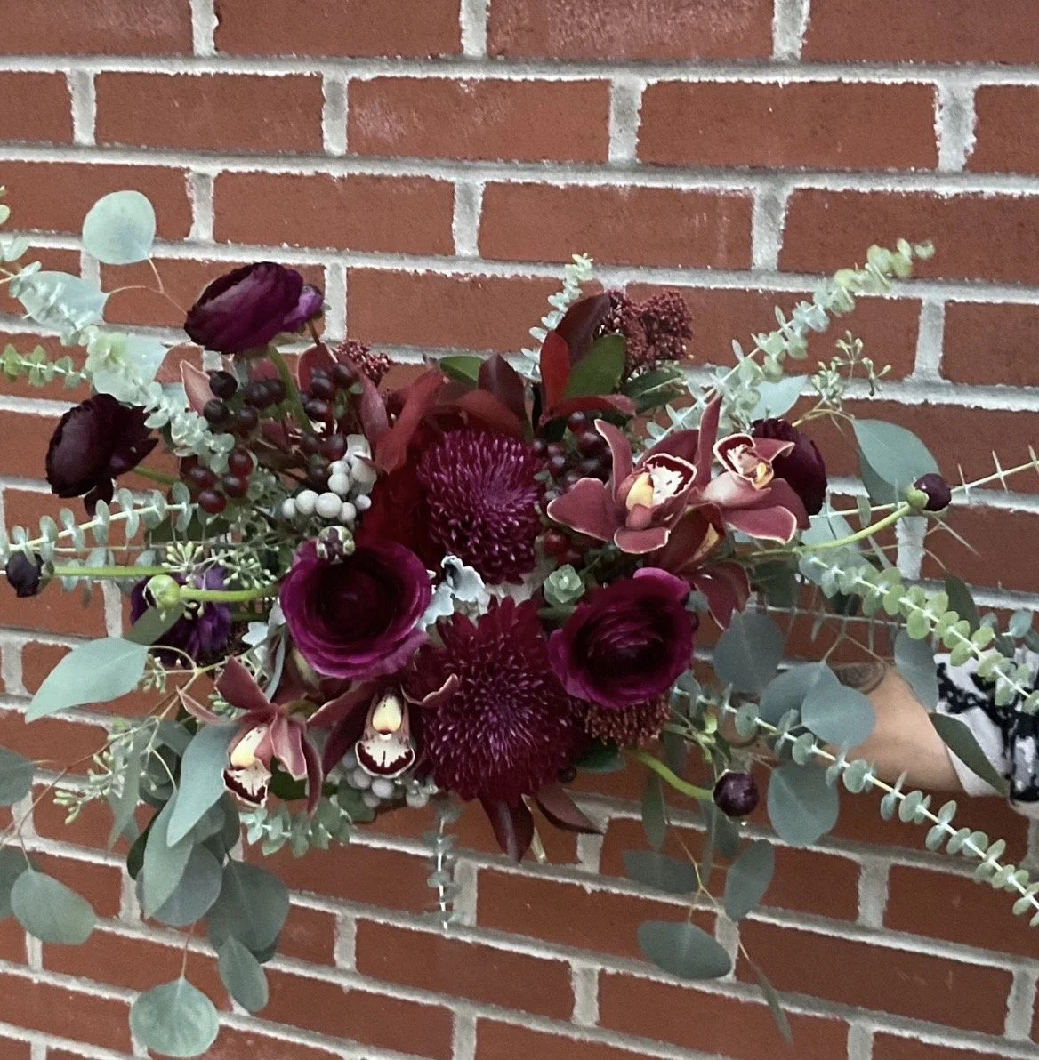 A floral bouquet featuring deep red and purple flowers, with greenery, held against a red brick wall.