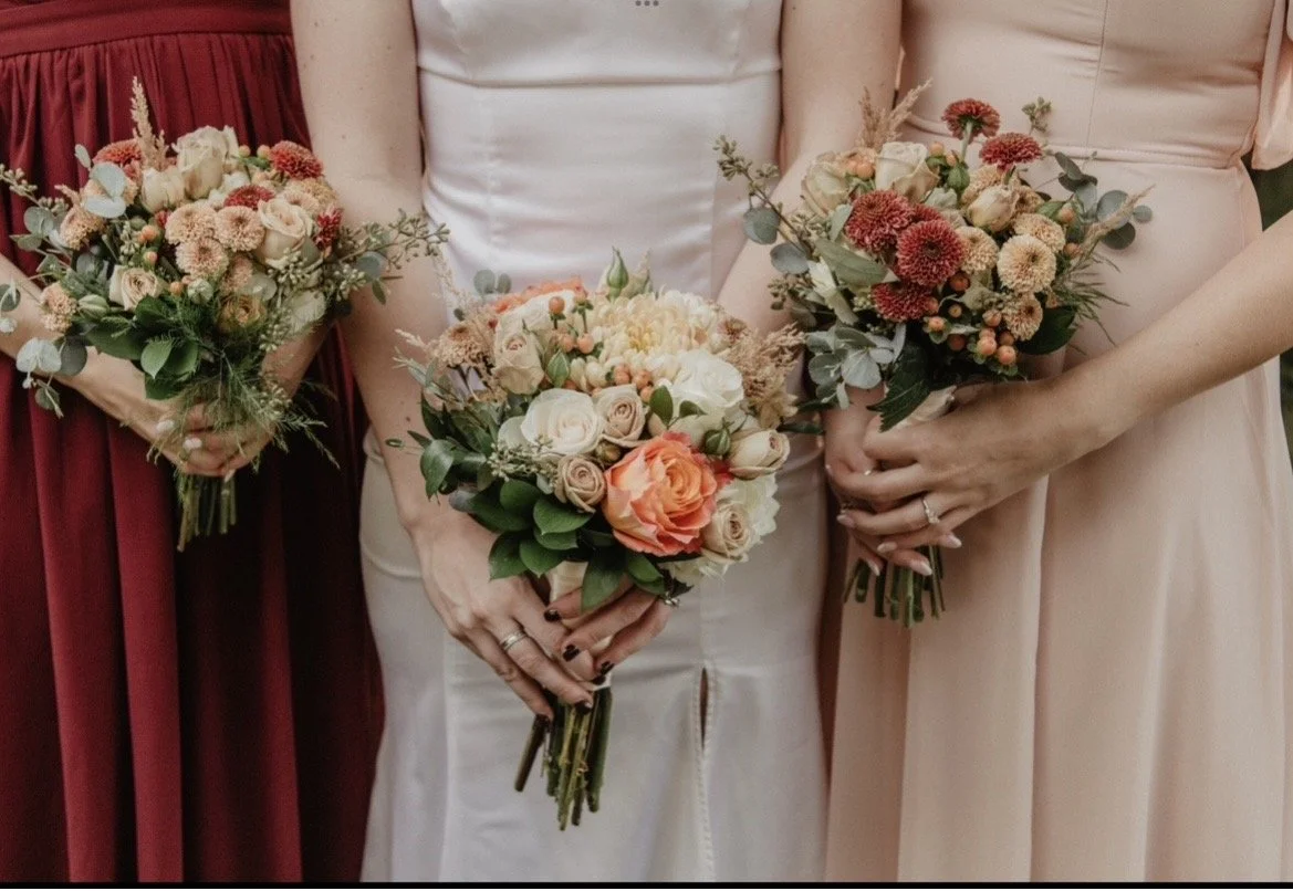Three women holding bouquets of flowers, with the middle woman wearing a white dress and the other two women wearing light pink and maroon dresses.