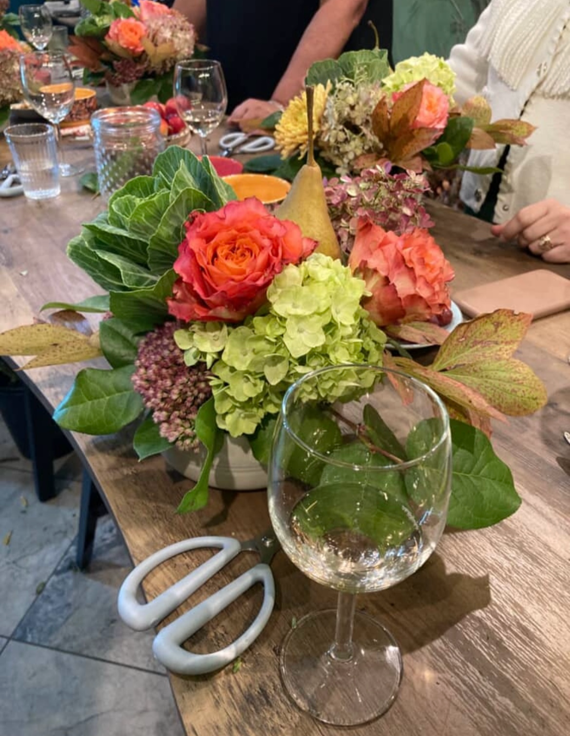 A floral centerpiece with pink roses, green hydrangeas, leaves, and other flowers on a wooden table, surrounded by glasses, plates, and other tableware at a gathering.
