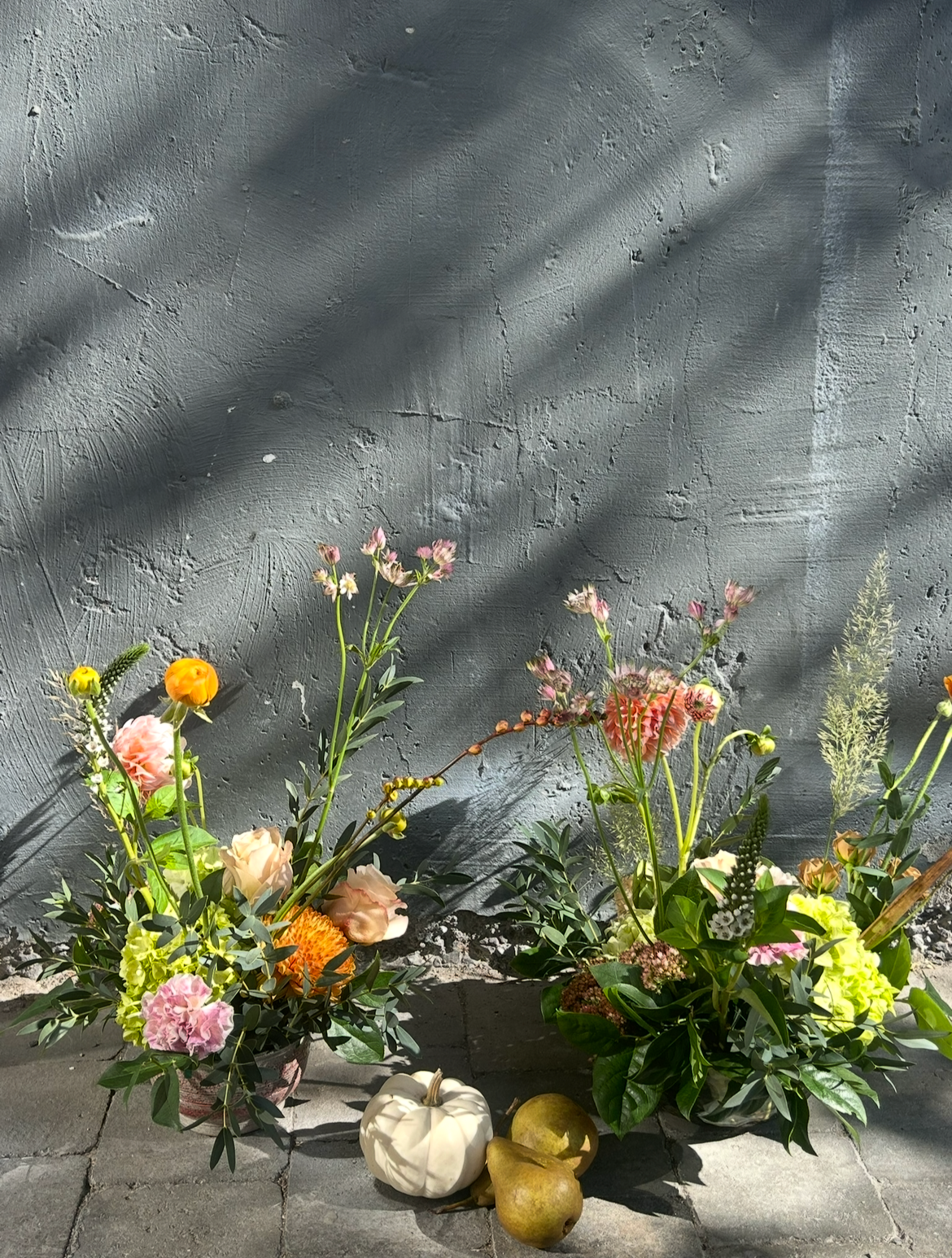Two floral arrangements with various colorful flowers, a white pumpkin, and two pears placed on a grey brick surface, set against a textured grey wall with shadows.