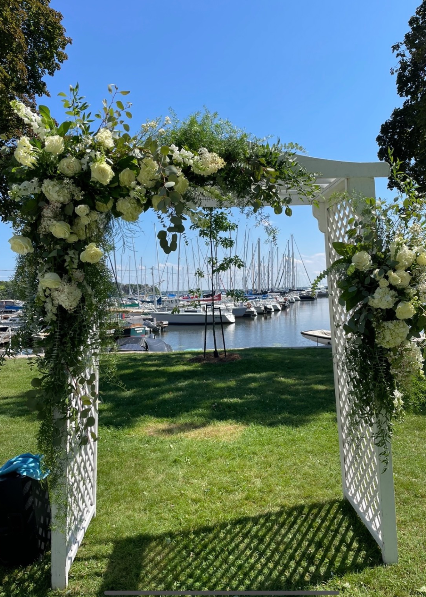 A white wooden wedding arch decorated with white flowers and greenery, set on a lawn near a marina with sailboats docked in water and a blue sky overhead.