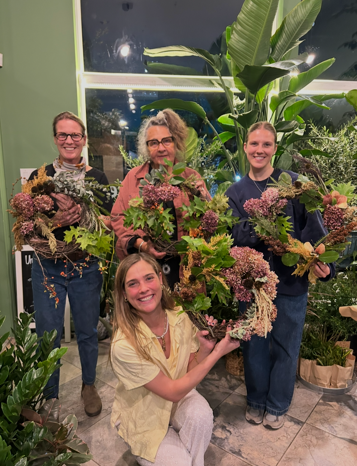 Four women holding floral arrangements inside a plant shop, with large green plants and potted plants around them.