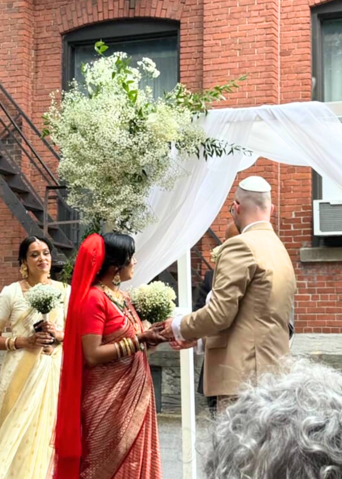 Jewish wedding ceremony outdoors with a bride in a red sari and a groom in a beige suit exchanging rings under a decorated arch with white fabric and flowers, in front of a red brick building.