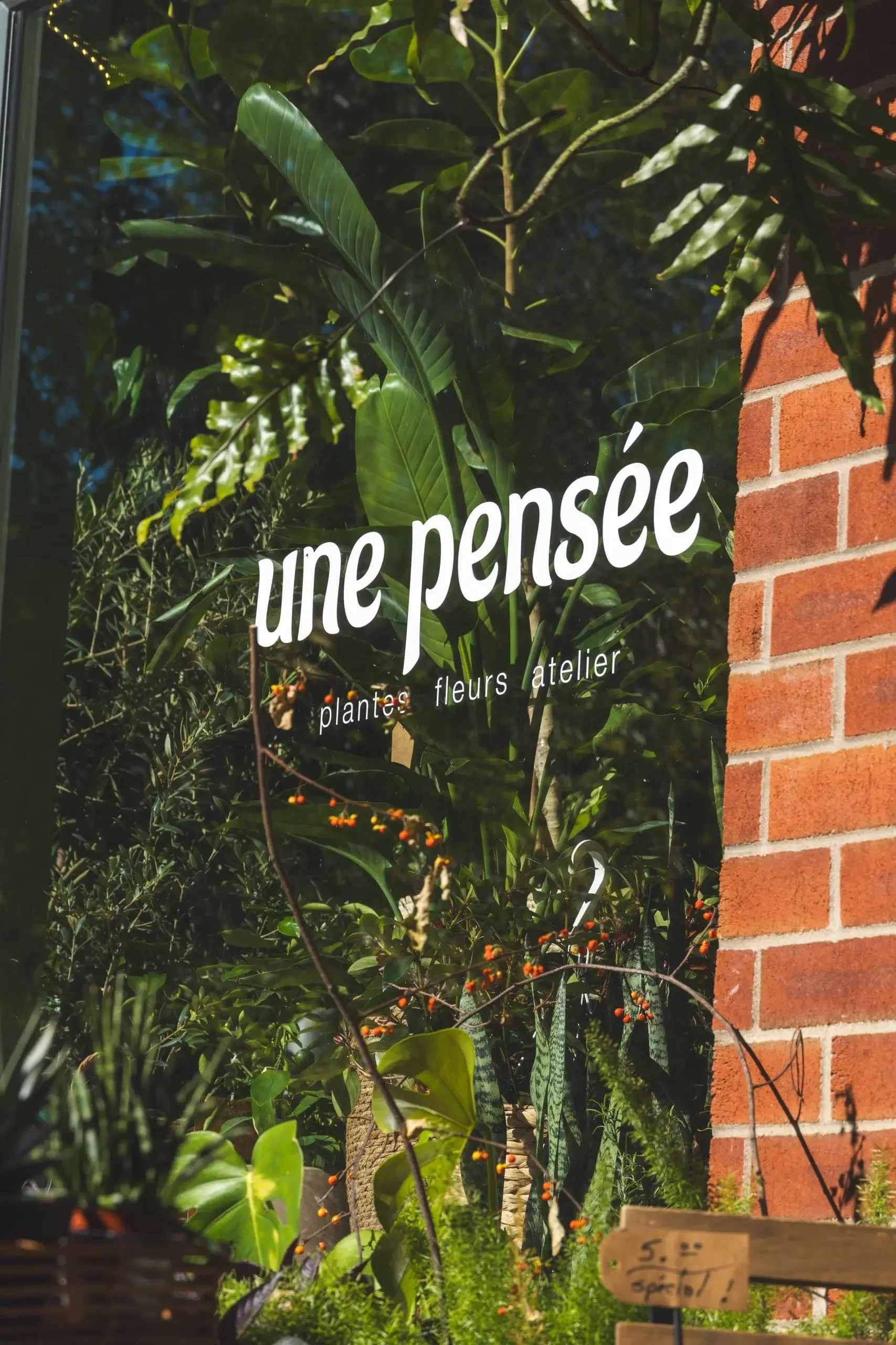 The image shows the exterior window of a plant shop called 'une pensée.' The shop's window displays the shop name and the words 'plantes, fleurs, atelier' written in white. Behind the window is lush green foliage, various plants, and a brick wall on 