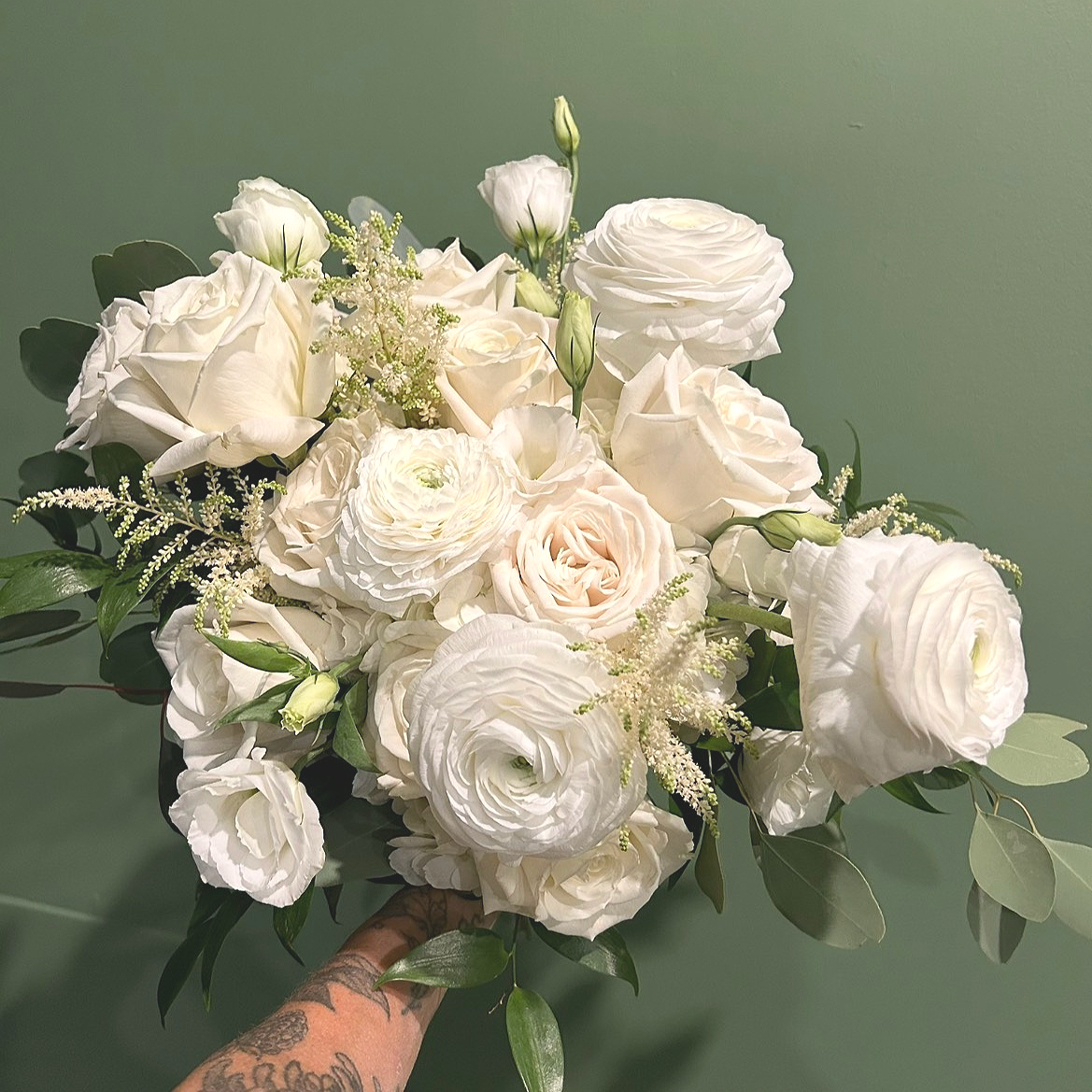 A bouquet of white roses, ranunculus, and delicate filler flowers with greenery, held by a tattooed hand, against a green background.