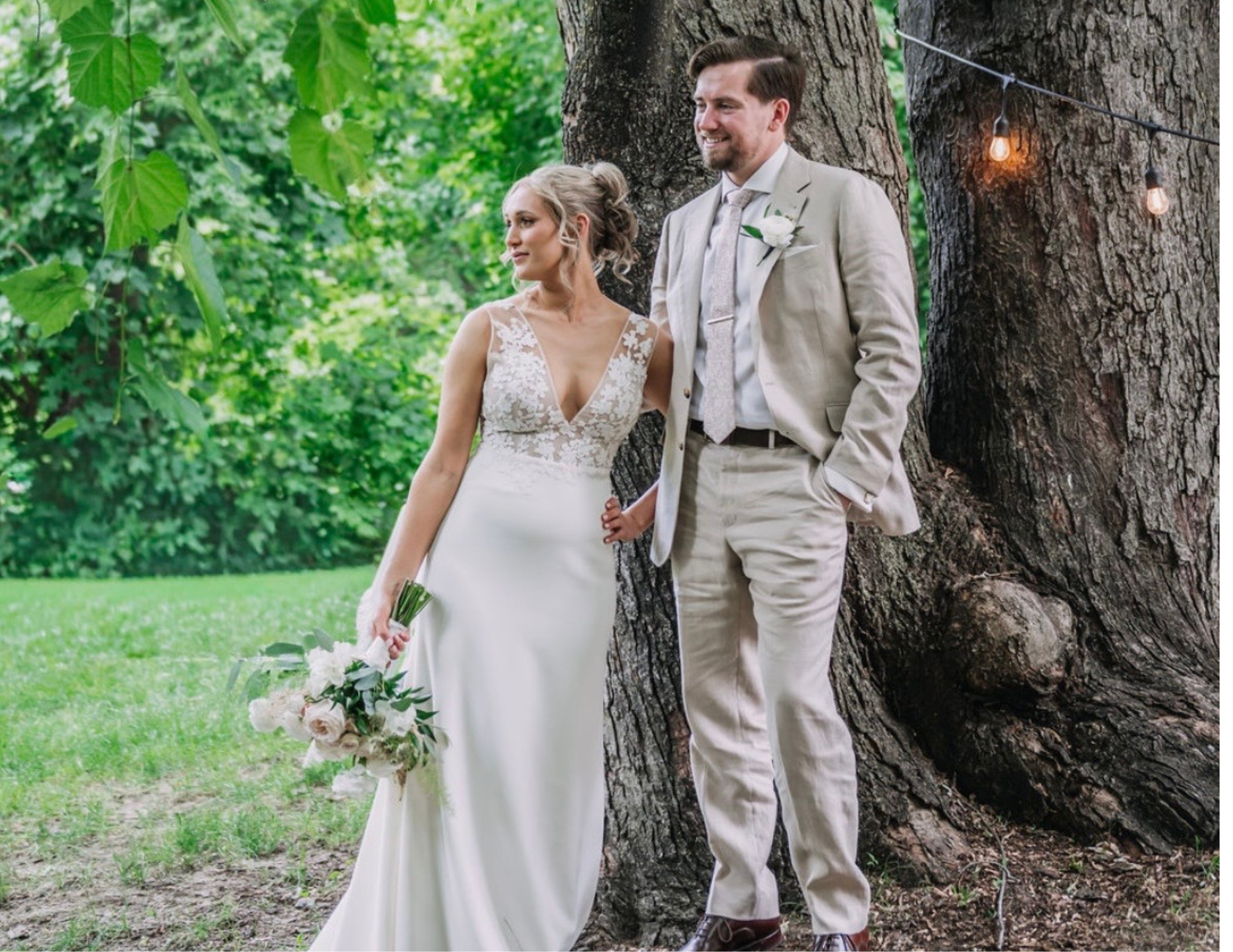 A bride and groom standing outdoors under a large tree, holding hands, with string lights hanging, greenery in the background, and the bride holding a bouquet of flowers.