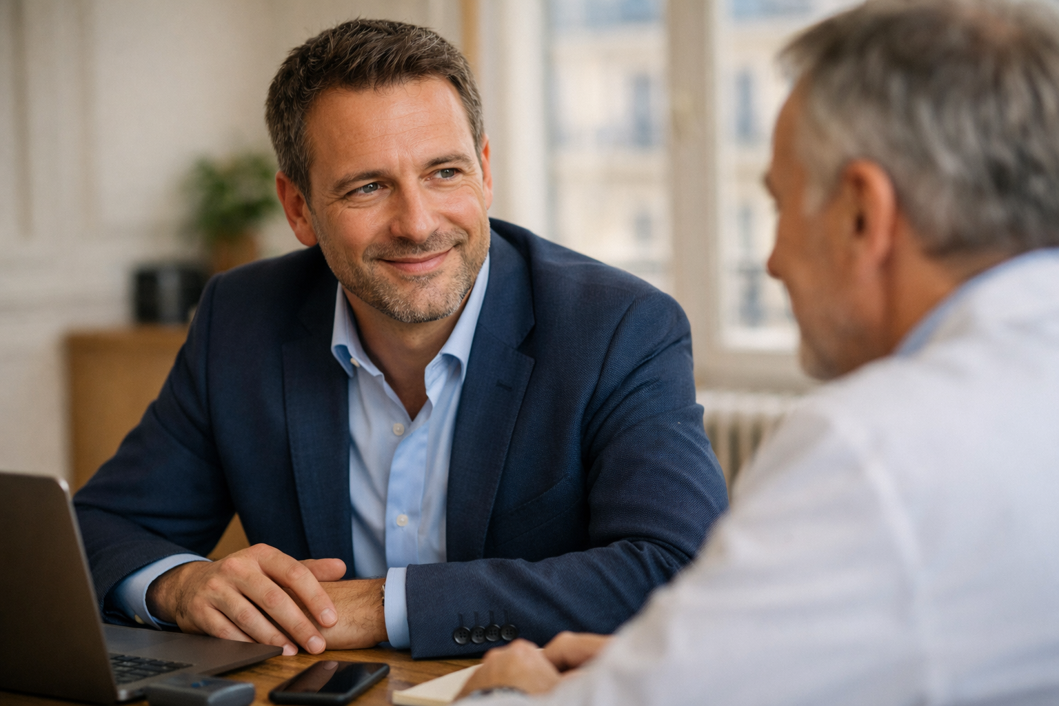 Deux hommes discutent dans un bureau. L'homme en premier plan porte une veste bleue et sourit. L'autre homme, de dos, porte une blouse blanche et écoute attentivement.