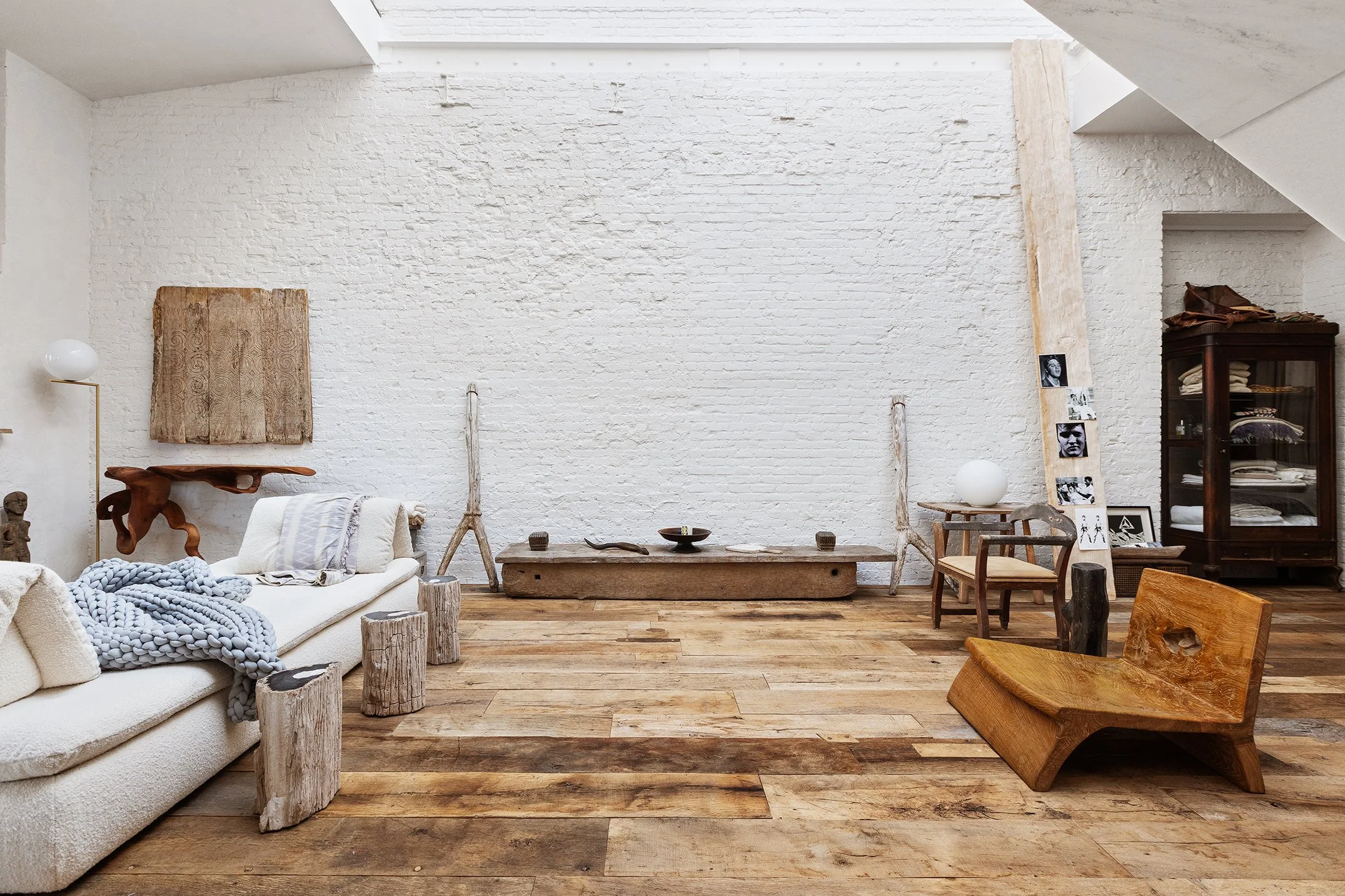 Minimalist living room with white brick wall, wooden floor, and eclectic furniture including a white sofa with throw blanket, wooden stools, a wooden bench, a glass display cabinet, and decorative items like photos, sculptures, and a lamp.