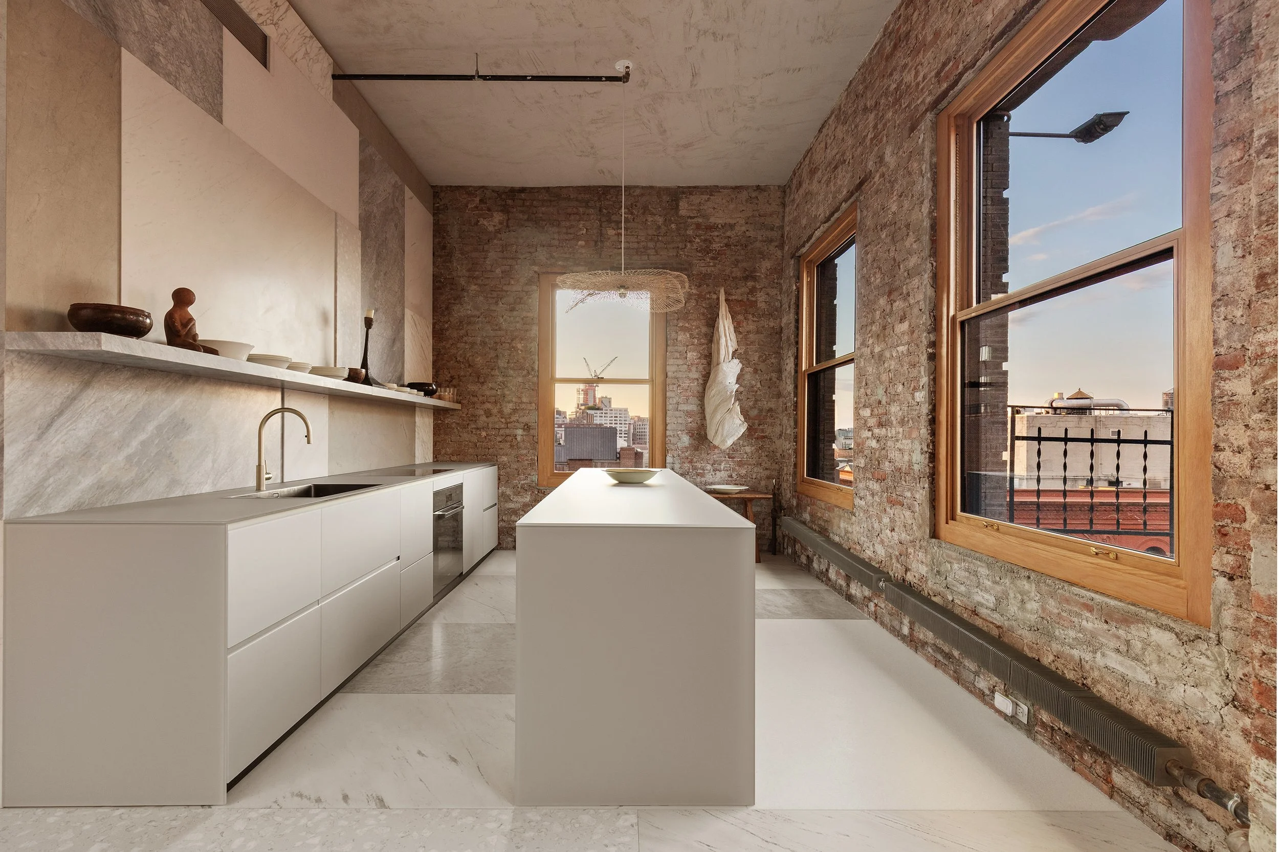 Modern kitchen with white cabinets, a beige marble countertop and backsplash, exposed brick walls, large windows letting in natural light, and an island in the center with a white bowl on top.