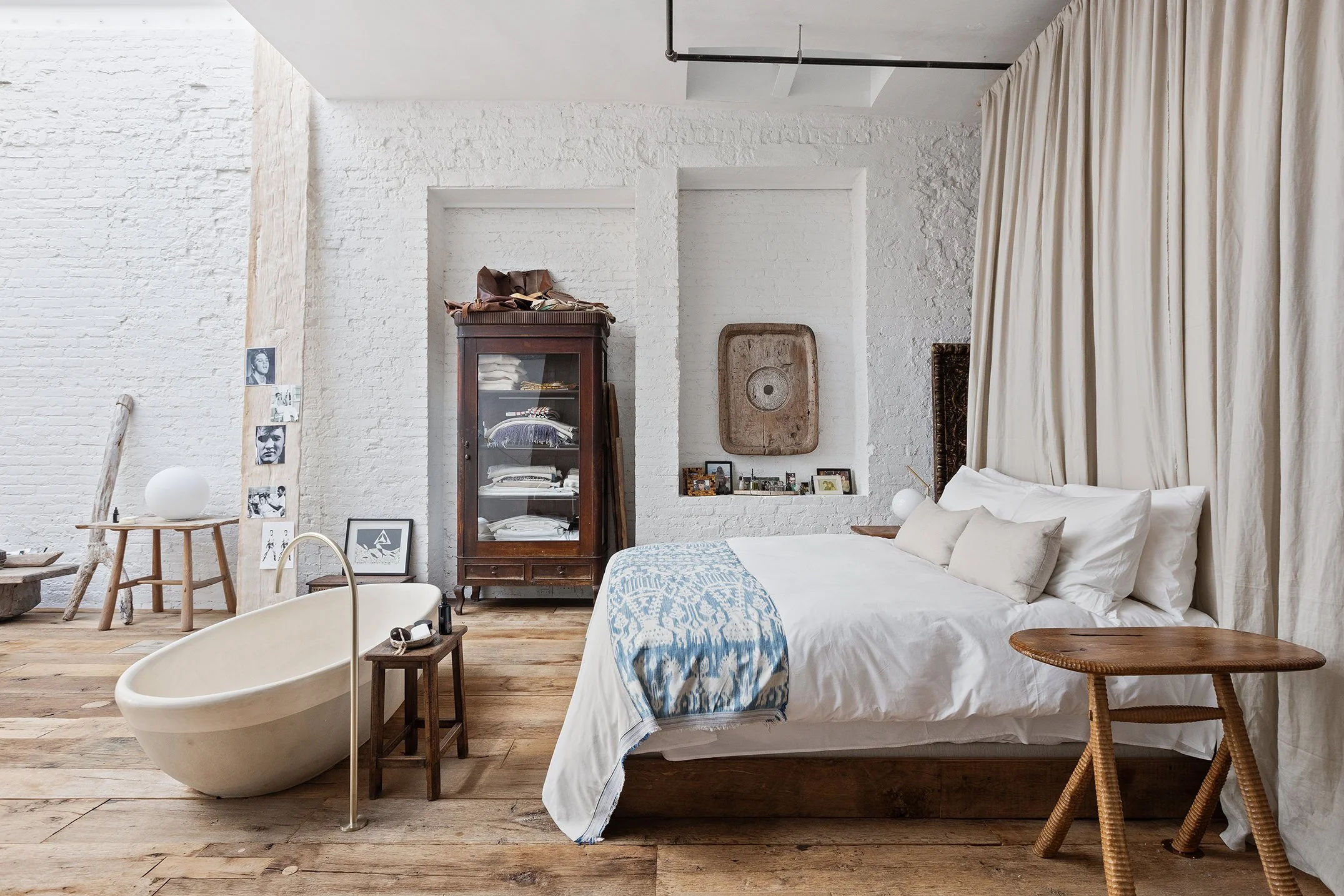 A minimalist bedroom with a white brick wall, wooden flooring, and a bed with white bedding and beige pillows. Next to the bed is a round wooden side table, and a large cream-colored curtain drapes behind the bed. There is a vintage wooden cabinet wi