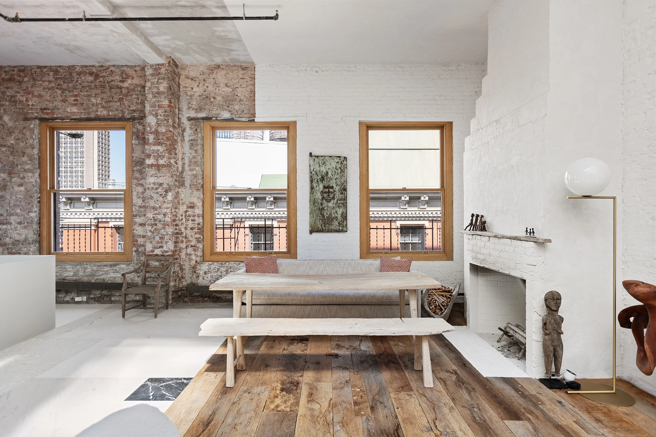 Interior of a loft-style living space with white and exposed brick walls, three wooden-framed windows, a fireplace with decorative objects, wooden flooring, and minimalist furniture, including a bench and a table.