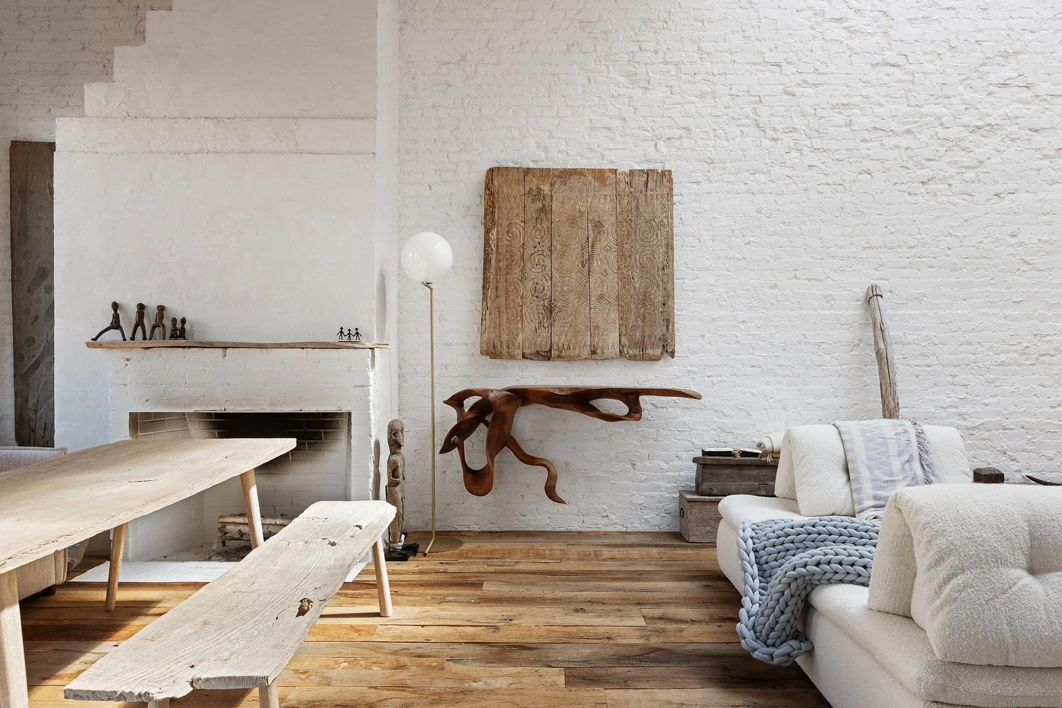 A cozy living room with white brick walls and wooden flooring, featuring a white couch with a chunky gray blanket, a rustic wooden table and bench, decorative sculptures, a tall white floor lamp, a hanging wooden art piece, and a fireplace mantle wit