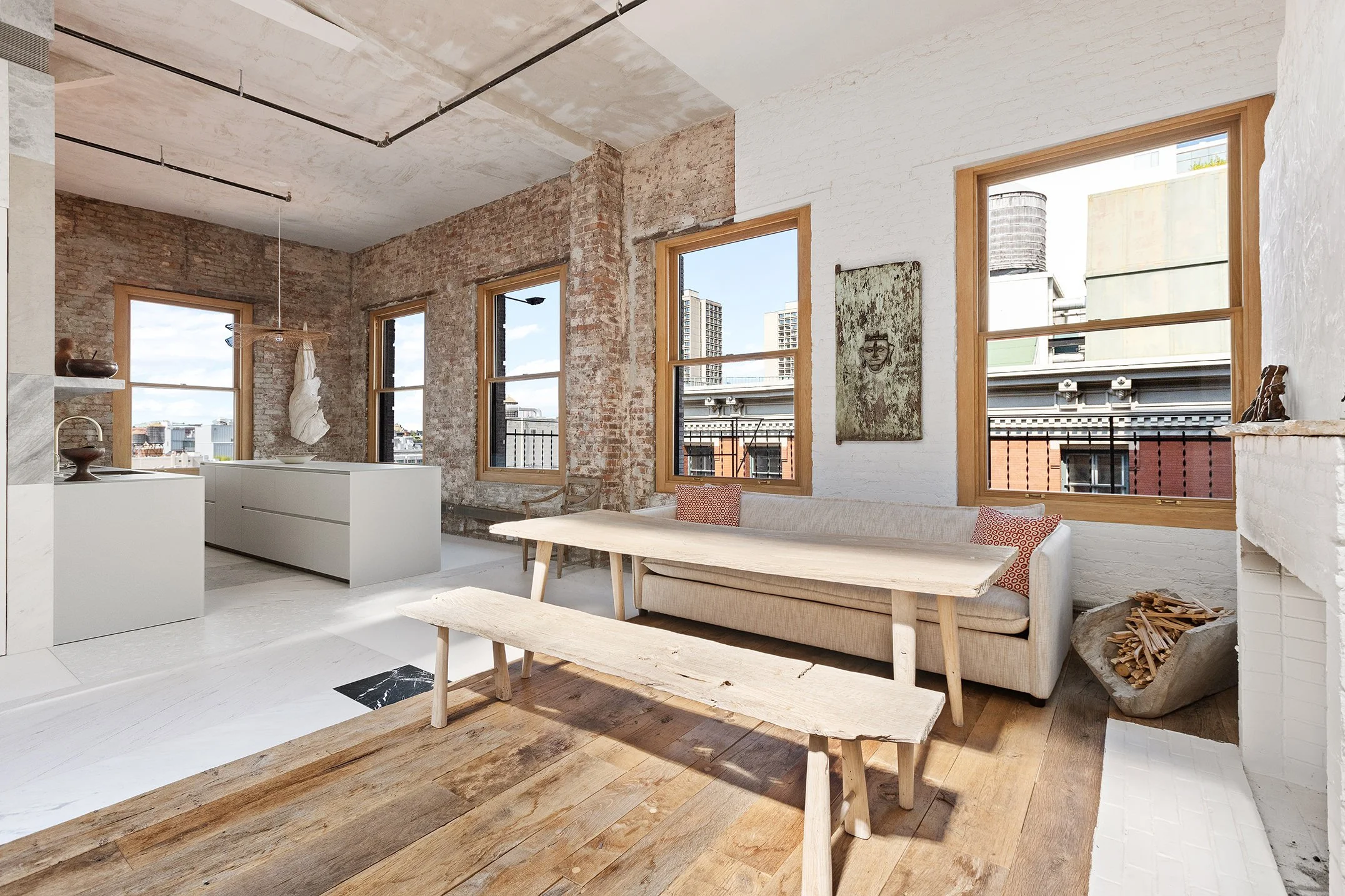 Loft-style interior living space with large windows, exposed brick walls, white walls, wooden furniture, a beige sofa with red pillows, a wooden bench, and a fireplace with firewood.