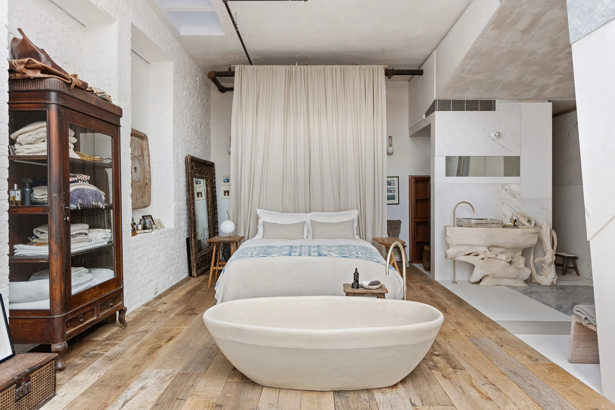 A bedroom with a white bed, light-colored bedding, and a sheer curtain behind the headboard. There's an oval-shaped bathtub in the foreground, wooden flooring, and a large mirror leaning against the wall to the left of the bed. A wooden cabinet is on