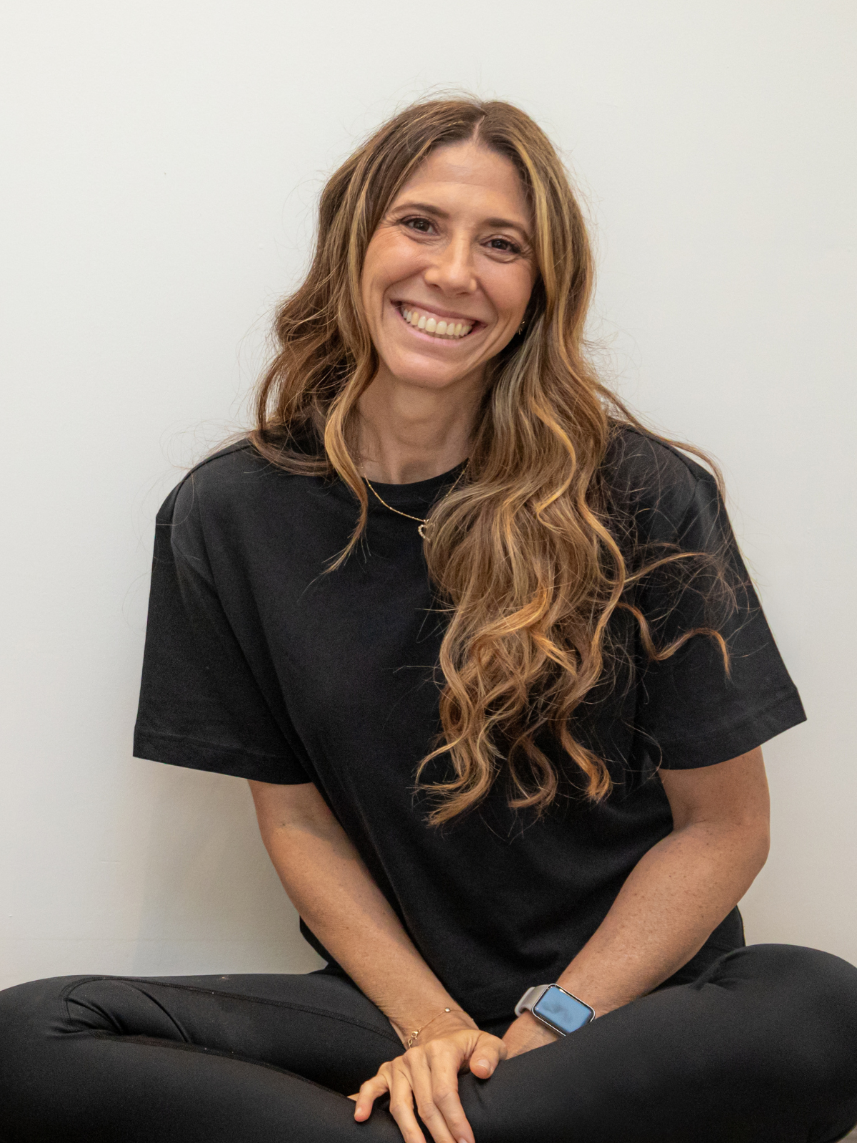 A woman with long wavy brown hair, wearing a black t-shirt and black pants, sitting cross-legged on the floor, smiling at the camera, against a plain white wall.