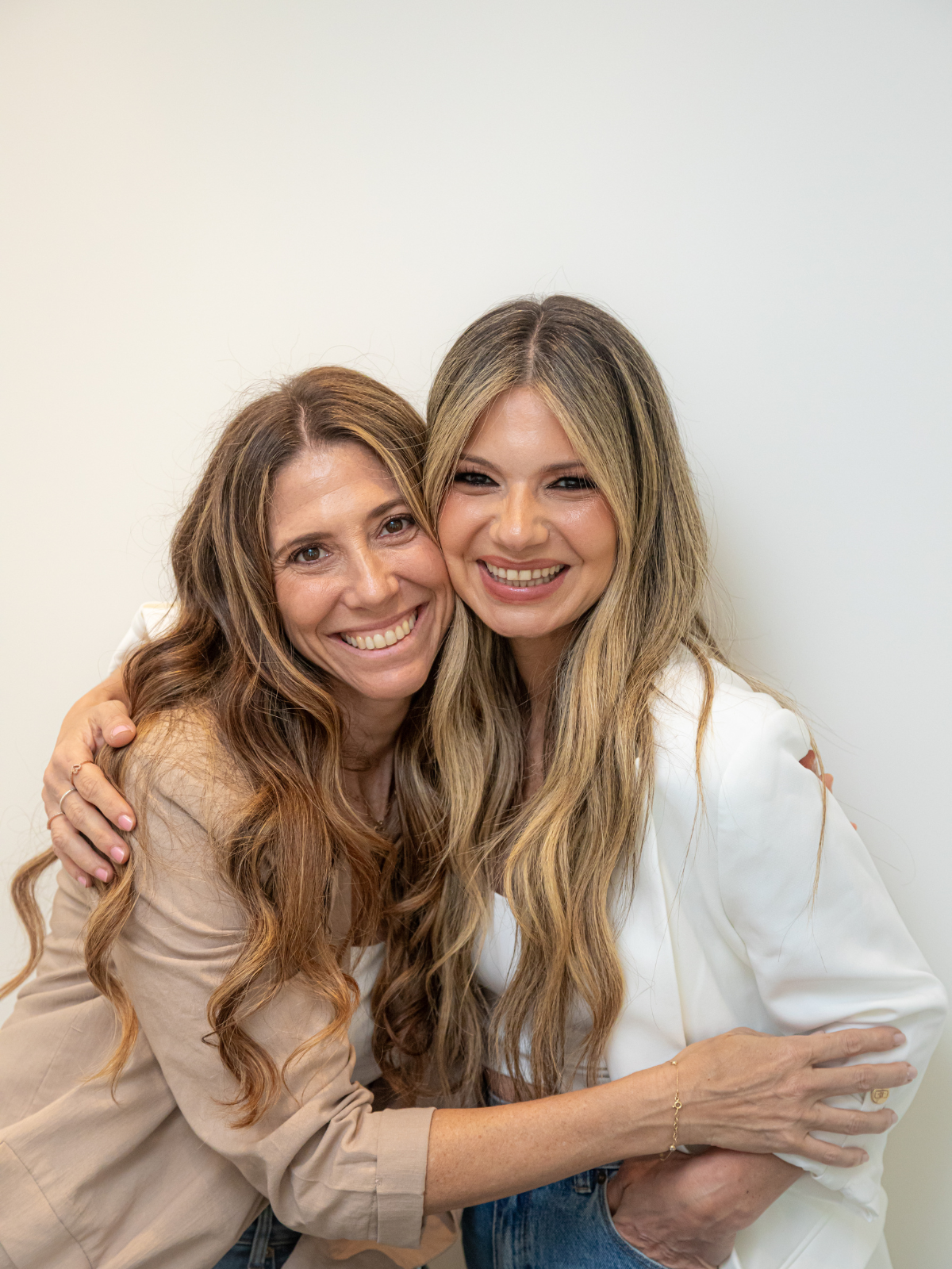 Two women with long wavy hair smiling and hugging each other, standing against a plain white wall.