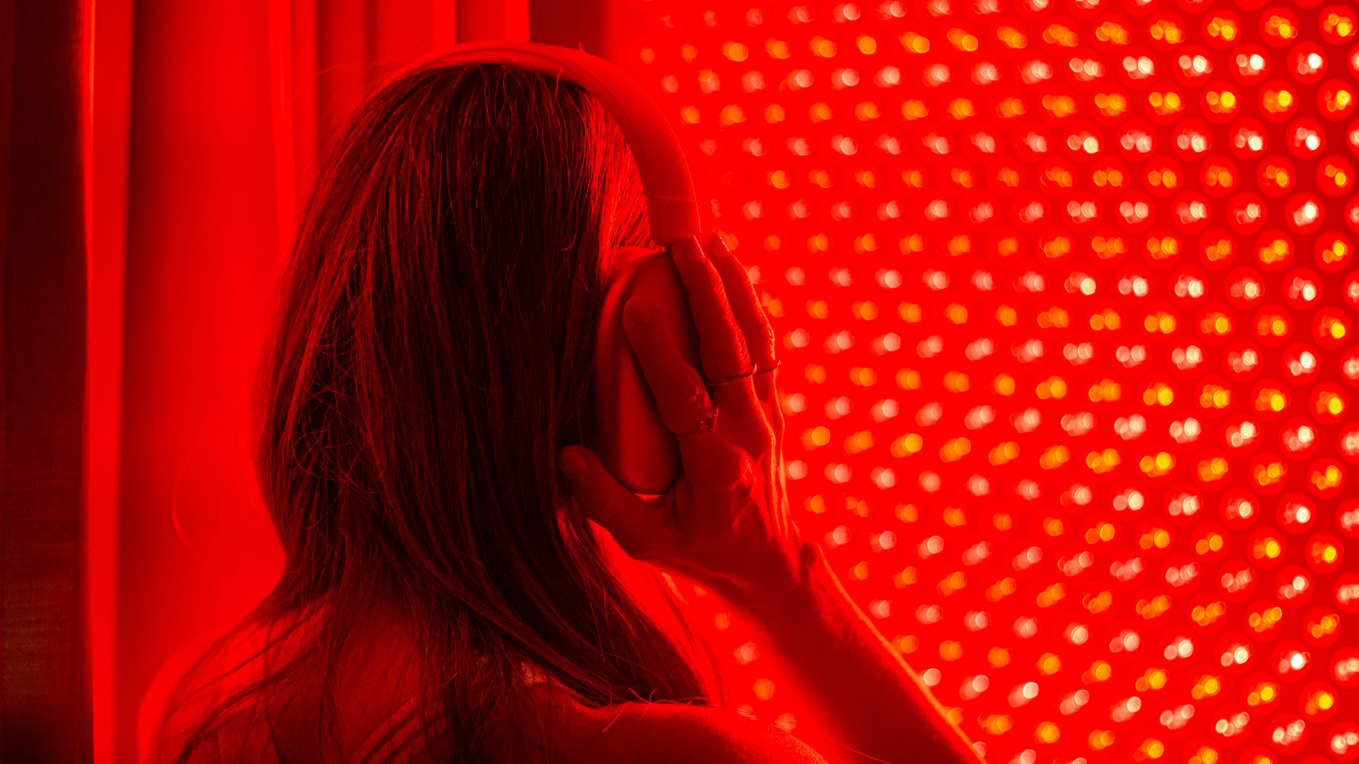 A person with long hair wearing headphones is shown in profile, surrounded by red lighting and small, bright red and yellow light spots in the background.