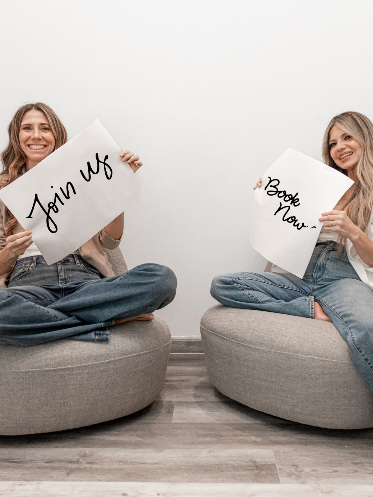 Two women sitting cross-legged on round ottomans, holding signs that say "Join Us" and "Book Now!" in front of a plain white wall, smiling at the camera.