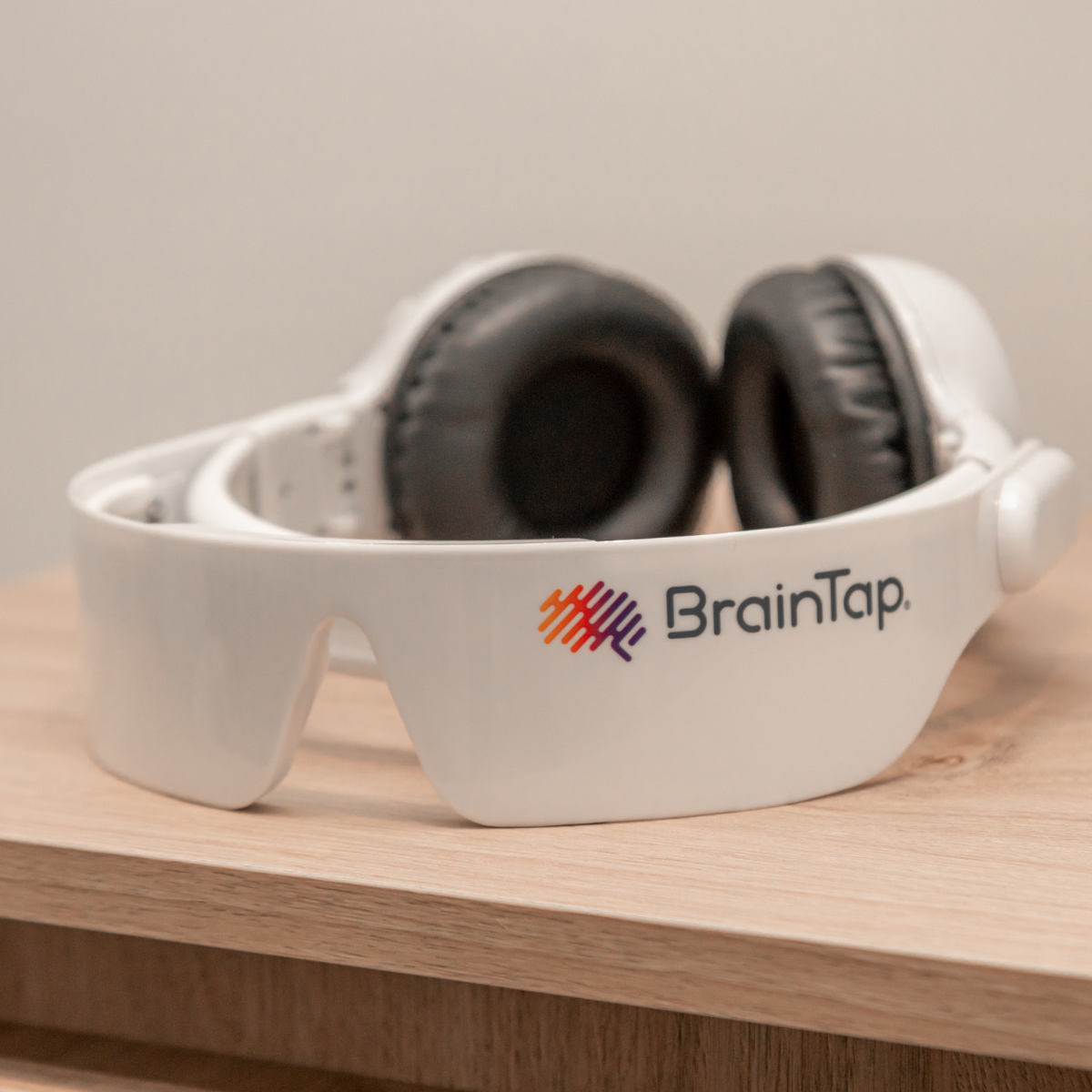 White BrainTap virtual reality headset with black headphones resting inside, placed on a wooden surface against a plain wall.