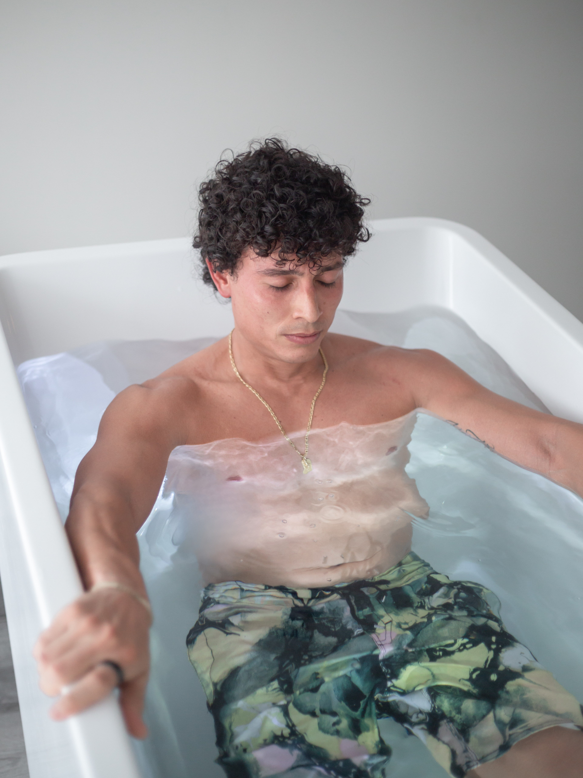 A man with curly dark hair relaxing in a white bathtub filled with water. He is wearing camouflage shorts and a gold chain necklace, with eyes closed and arms resting on the sides of the tub.