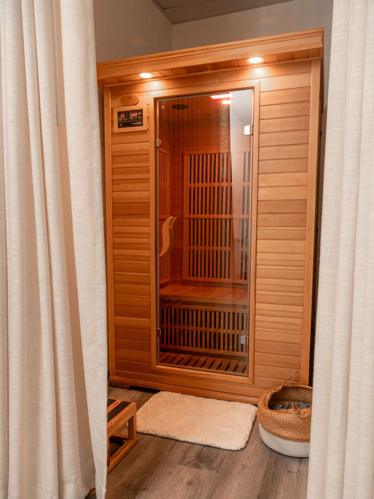 A wooden infrared sauna with a glass door. The sauna has a digital control panel and interior wooden slats. It is situated in a room with beige curtains, a white plush rug, and a wicker basket with folded towels.