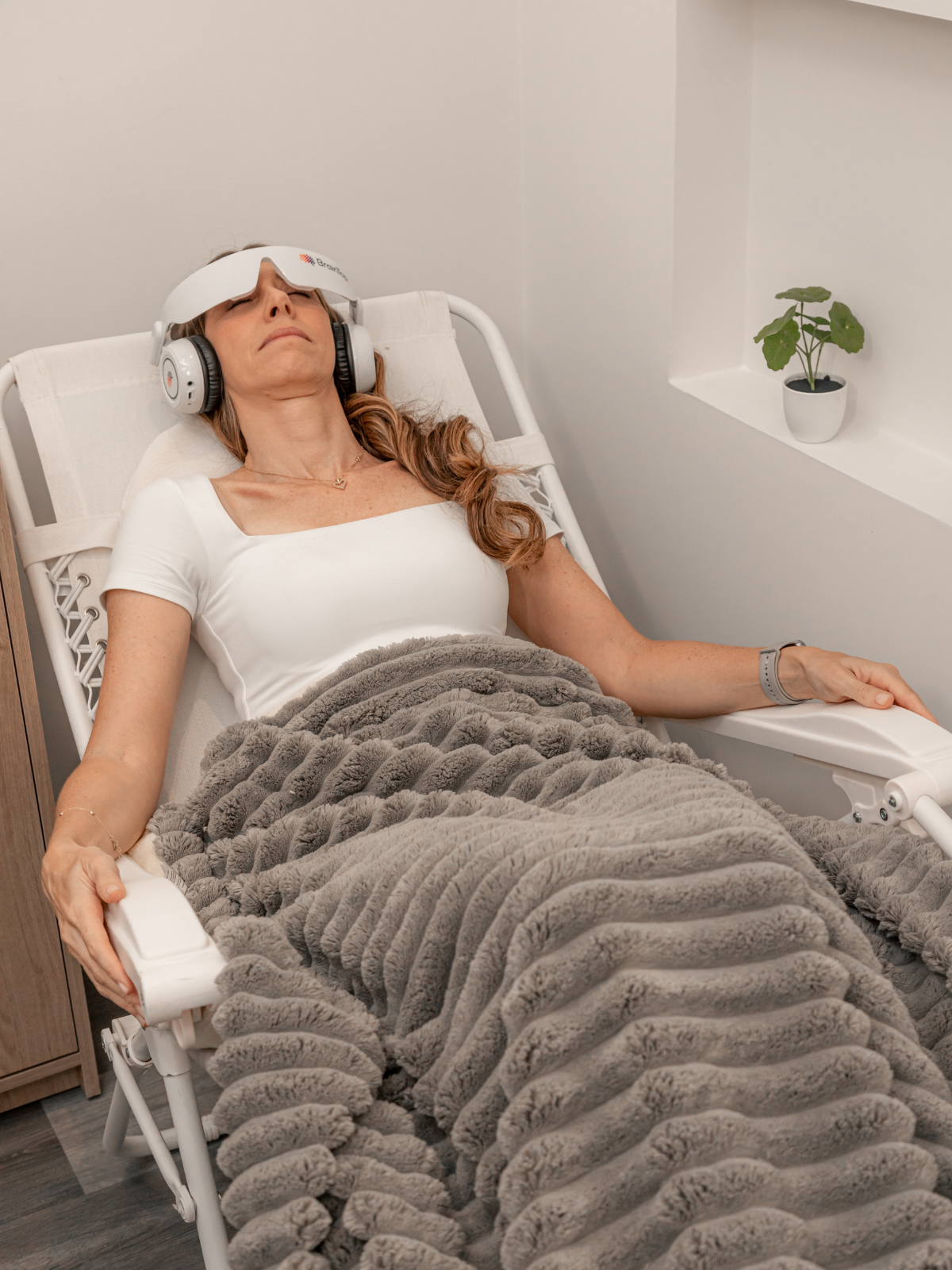 A woman lying on a medical recliner with a gray blanket, wearing a sleep mask and headphones, in a room with a small potted plant in a shelf.