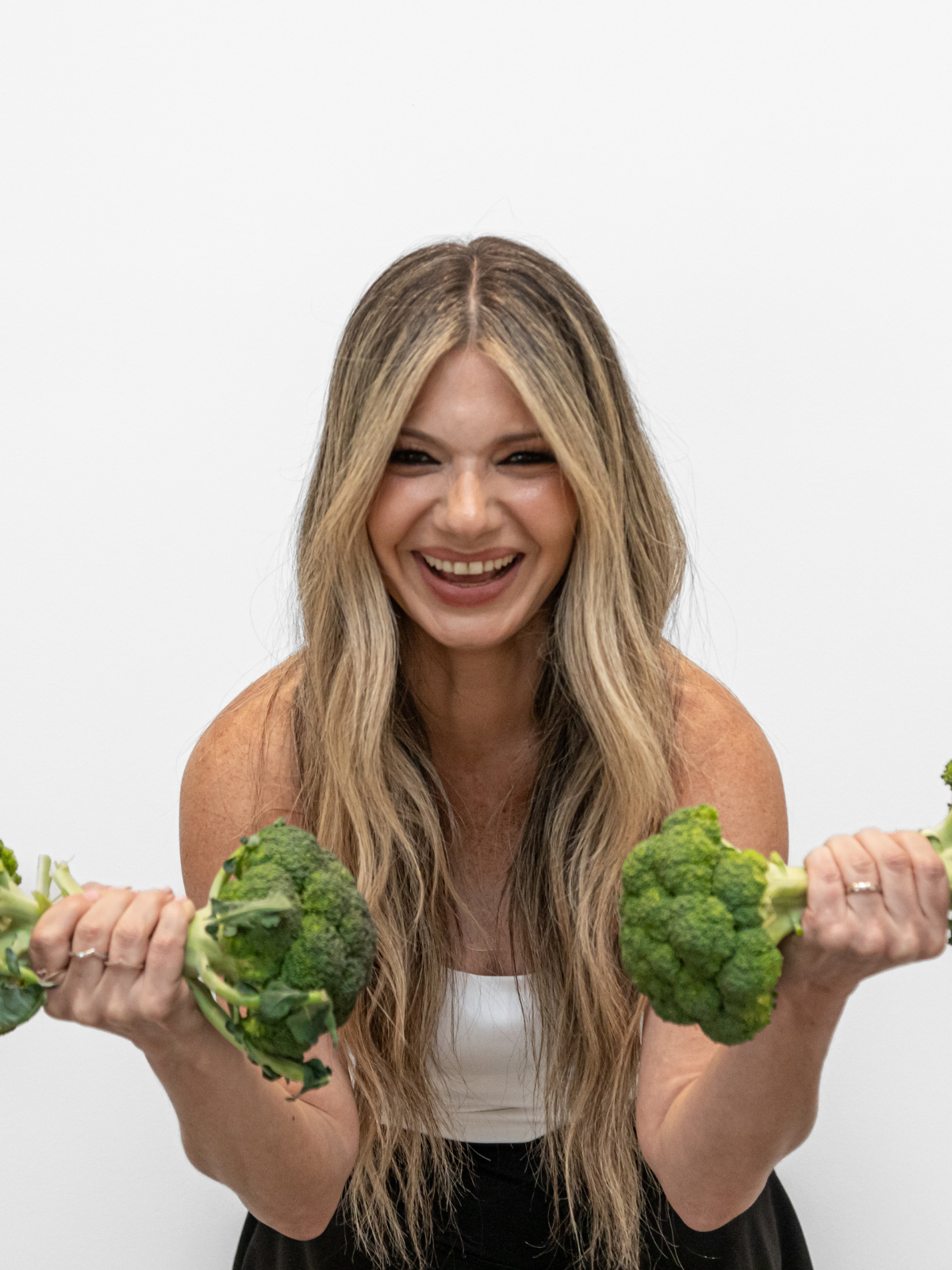 A woman with long, wavy blonde hair smiling and holding broccoli in both hands in front of a plain white background.