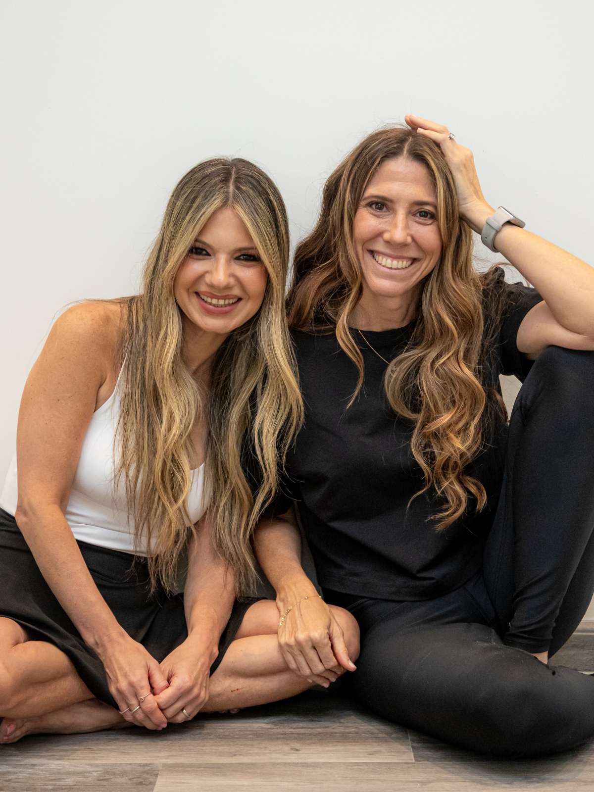 Two women sitting on the floor, smiling, with one woman touching her head, against a plain white background