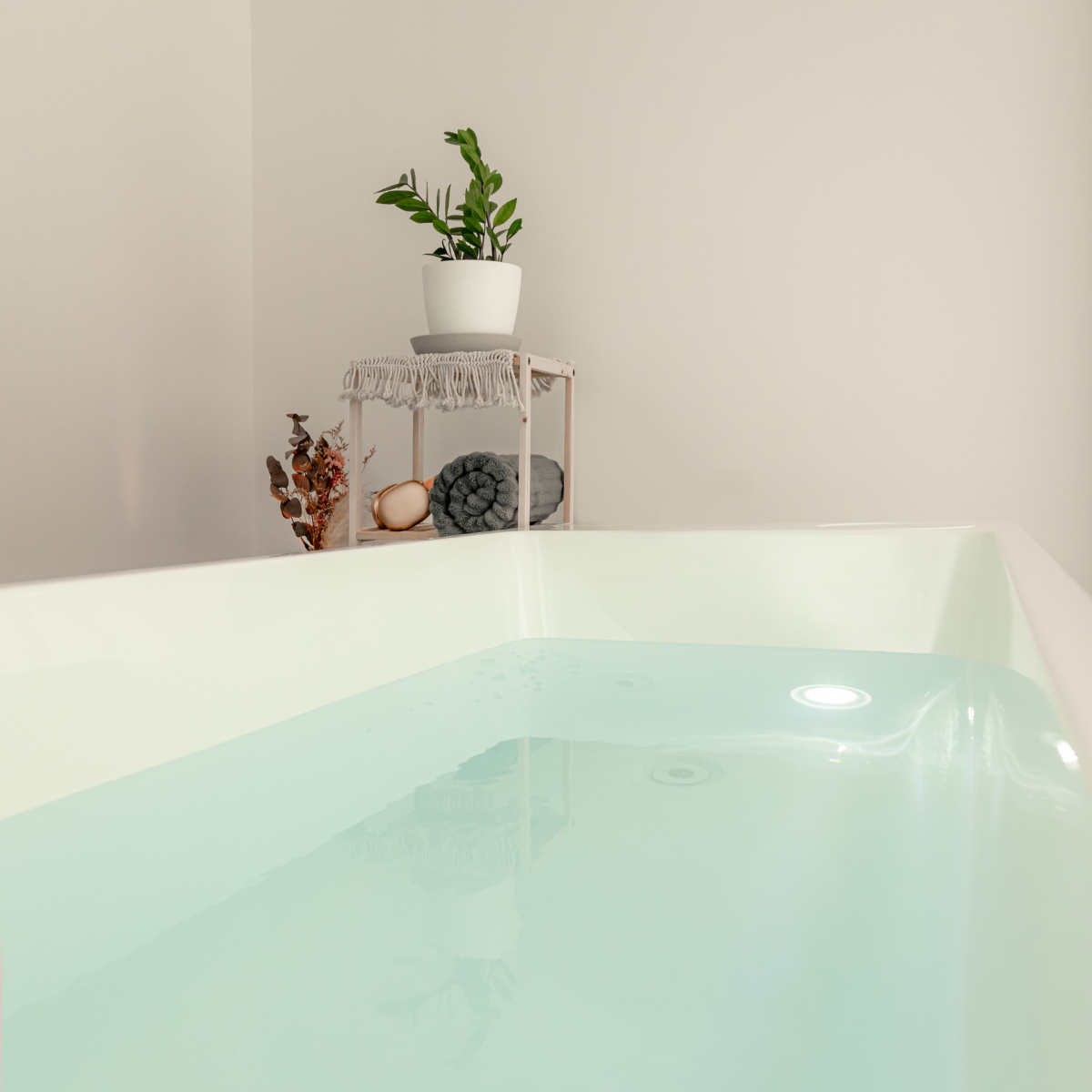 A light-colored bathtub filled with water in a bathroom with a white wall. In the background, there is a small wooden stand with a potted plant on top, and folded towels and decorative items on the lower shelf.