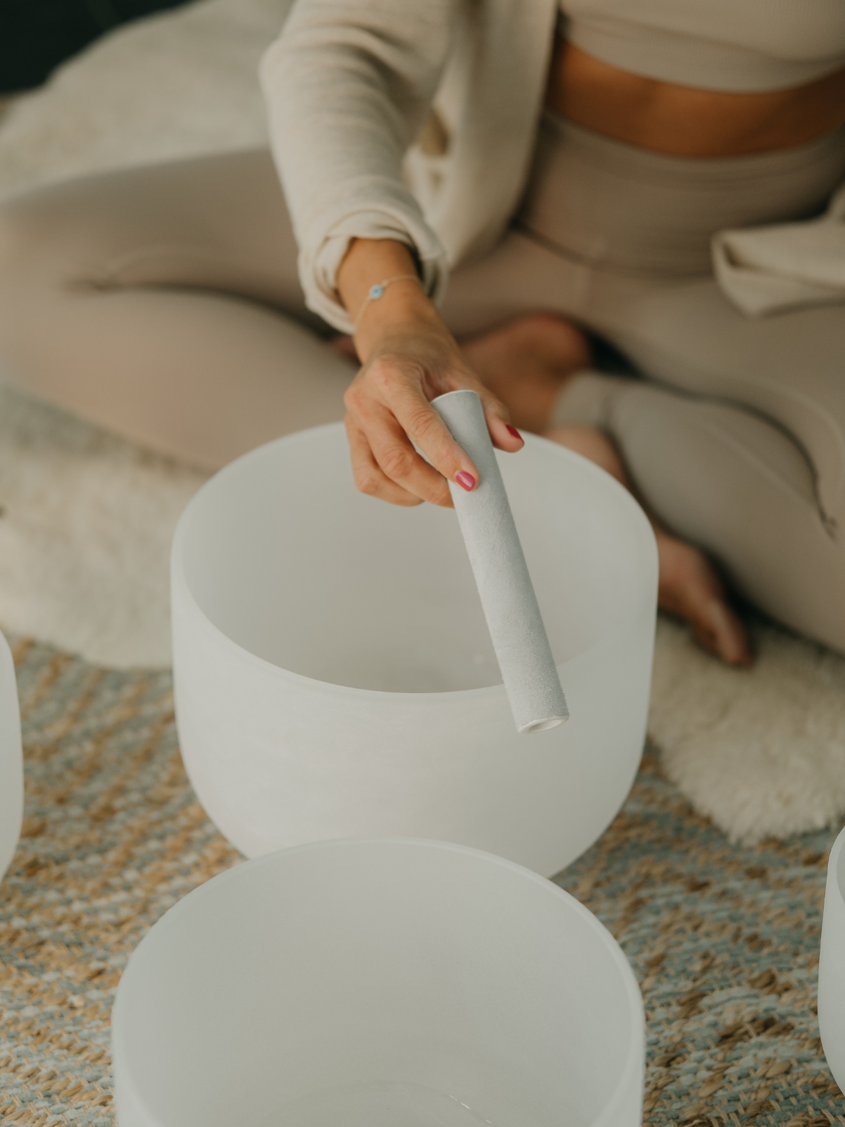 A woman is holding a white rod above a white bowl, with other similar bowls in front of her. She is seated on a patterned rug in a cozy room.