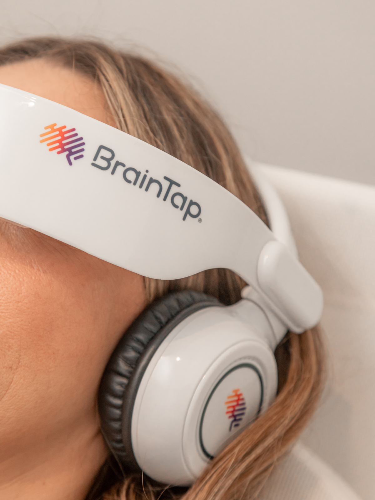 Close-up of a woman wearing BrainTap headphones with a white headband displaying the BrainTap logo, consisting of colorful lines and the brand name, in a clinical setting.