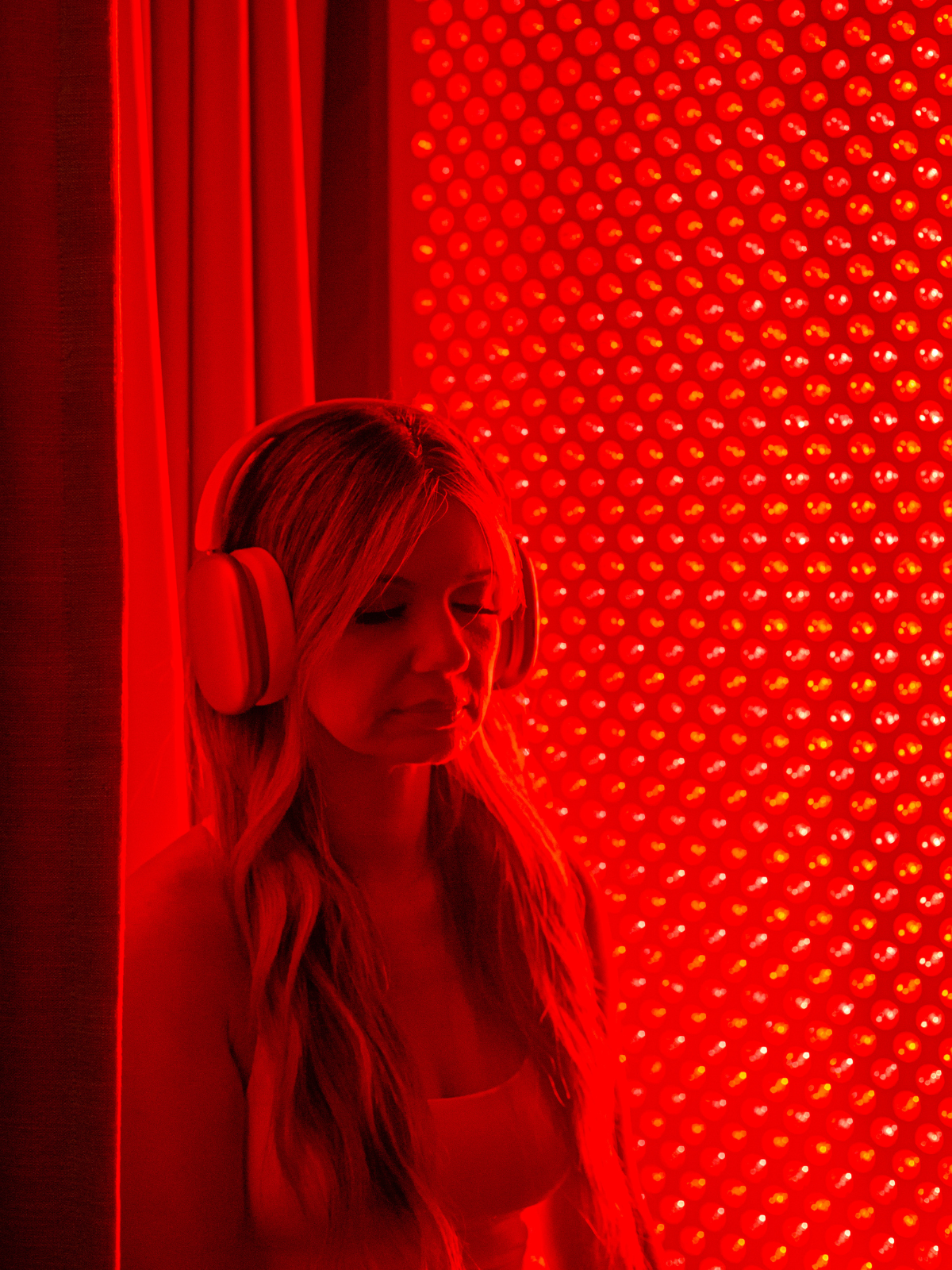 A woman with long hair wearing large headphones stands in front of a red-lit wall covered with bulbs, with her eyes closed.
