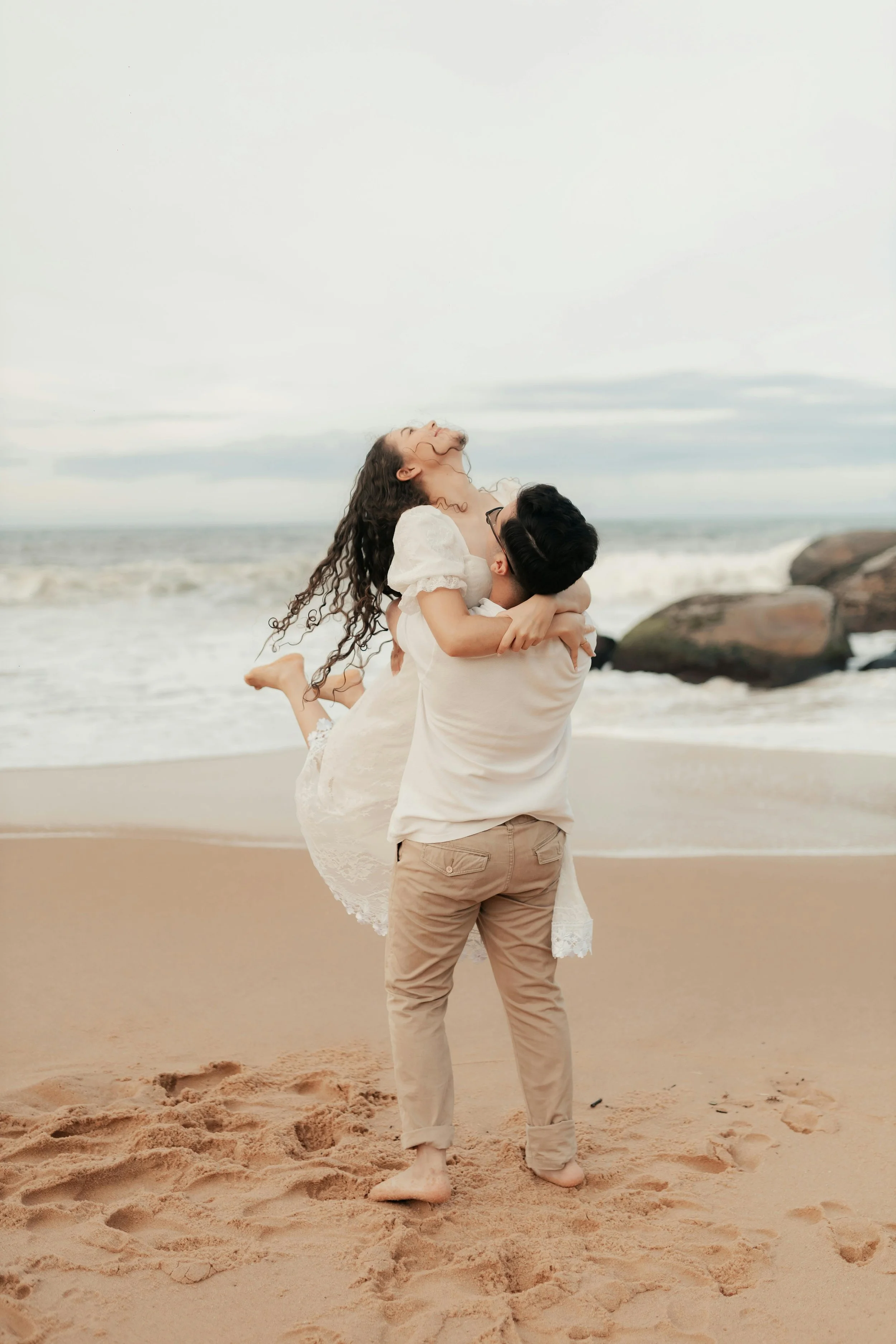 A man in khaki pants and a white shirt lifting a woman in a white dress on a sandy beach near rocks with waves in the background.