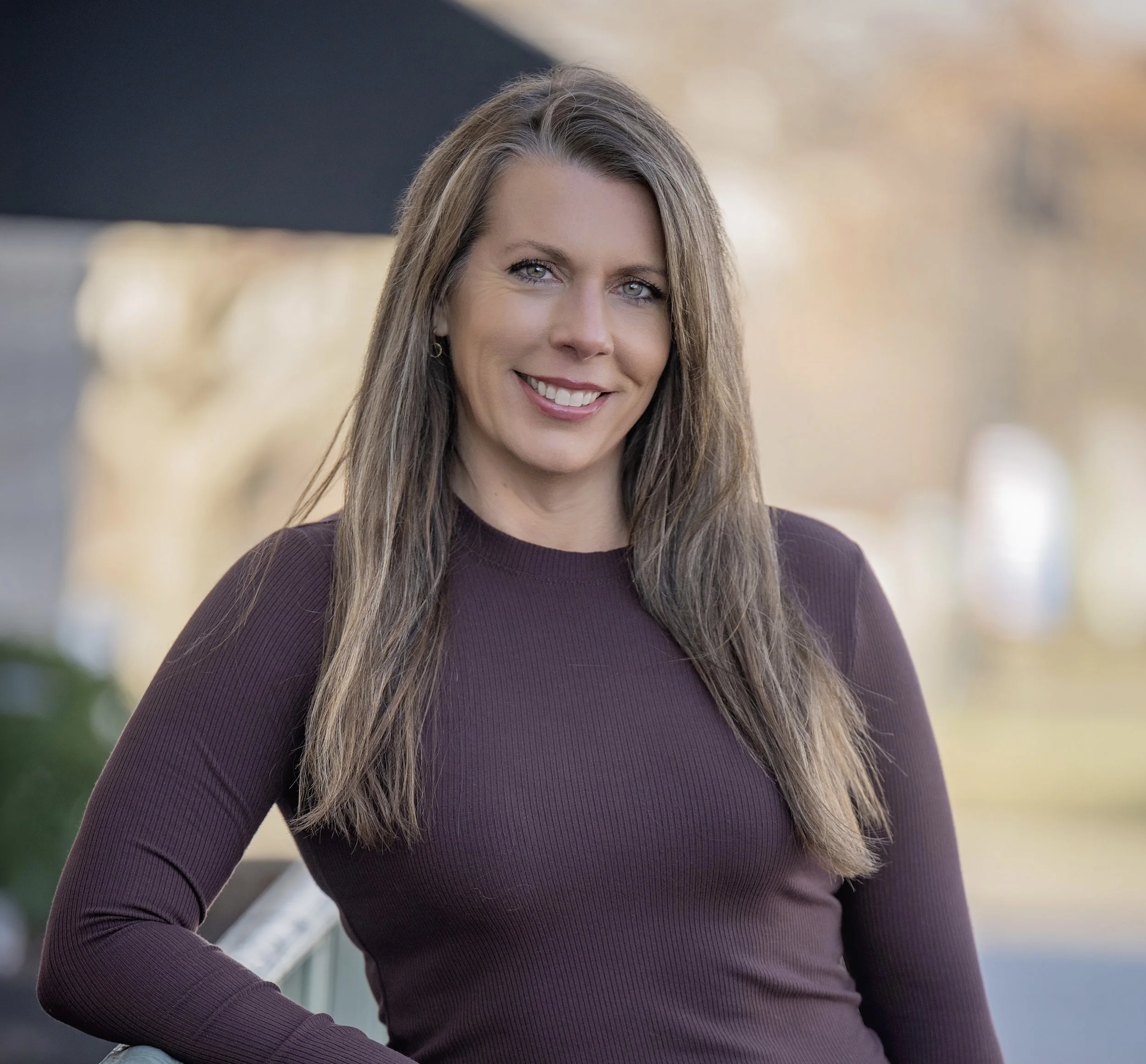 A woman with long brown hair and blue eyes smiling at the camera, wearing a maroon long-sleeve shirt, outdoors with a blurred natural background.