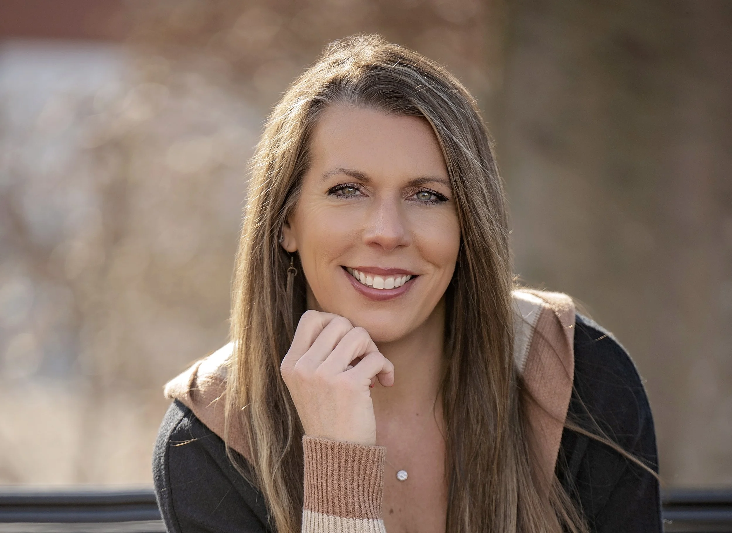 A smiling woman with long brown hair, wearing a black top and a brown hooded jacket, outdoors with a blurred background.