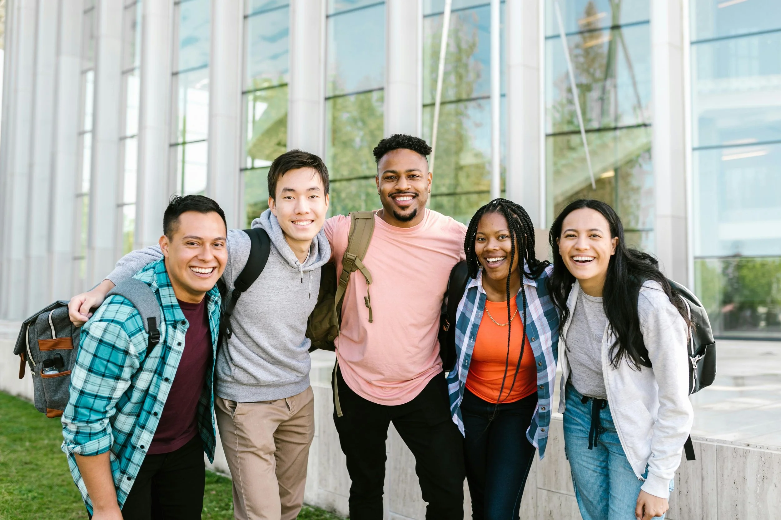 Group of five diverse young adults smiling with arms around each other outside in front of a modern glass building.