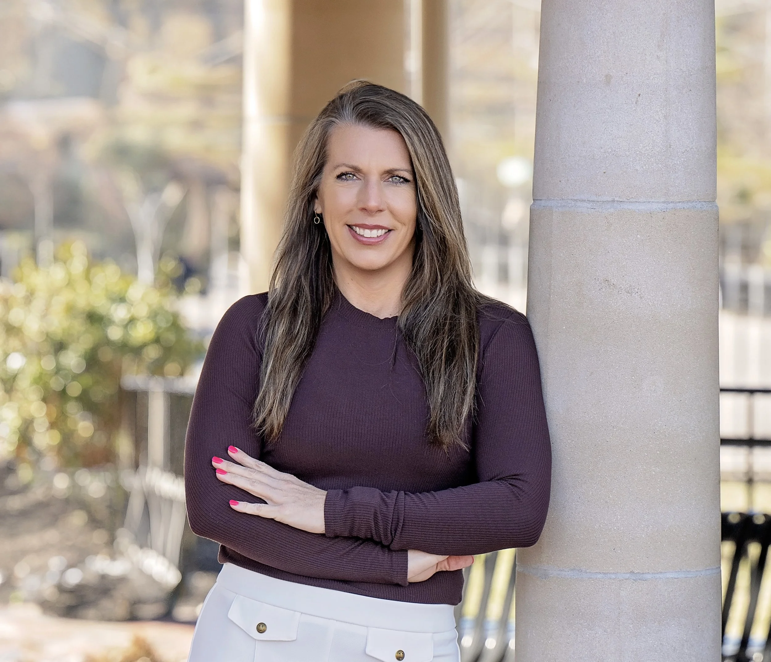 A smiling woman with shoulder-length brown hair, wearing a purple long-sleeve top and white pants, standing outdoors next to a concrete pillar in a park-like setting.